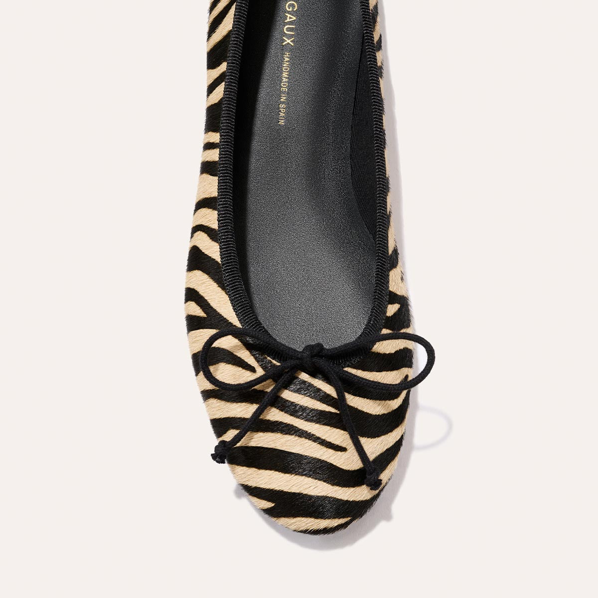 The Margaux Demi - Tan Zebra Haircalf is a handmade in Spain ballet flat featuring a tan and black zebra print, a rounded toe, and a small black front bow.