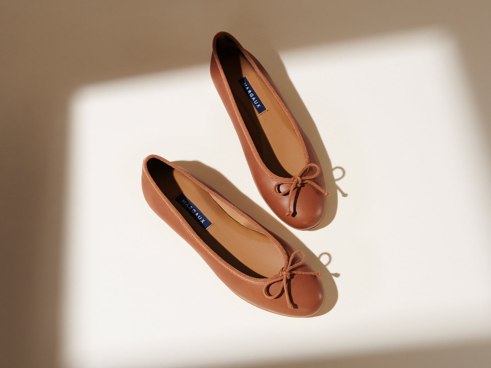 A pair of brown ballet flats with bows on the toes, placed on a light surface with soft shadows.