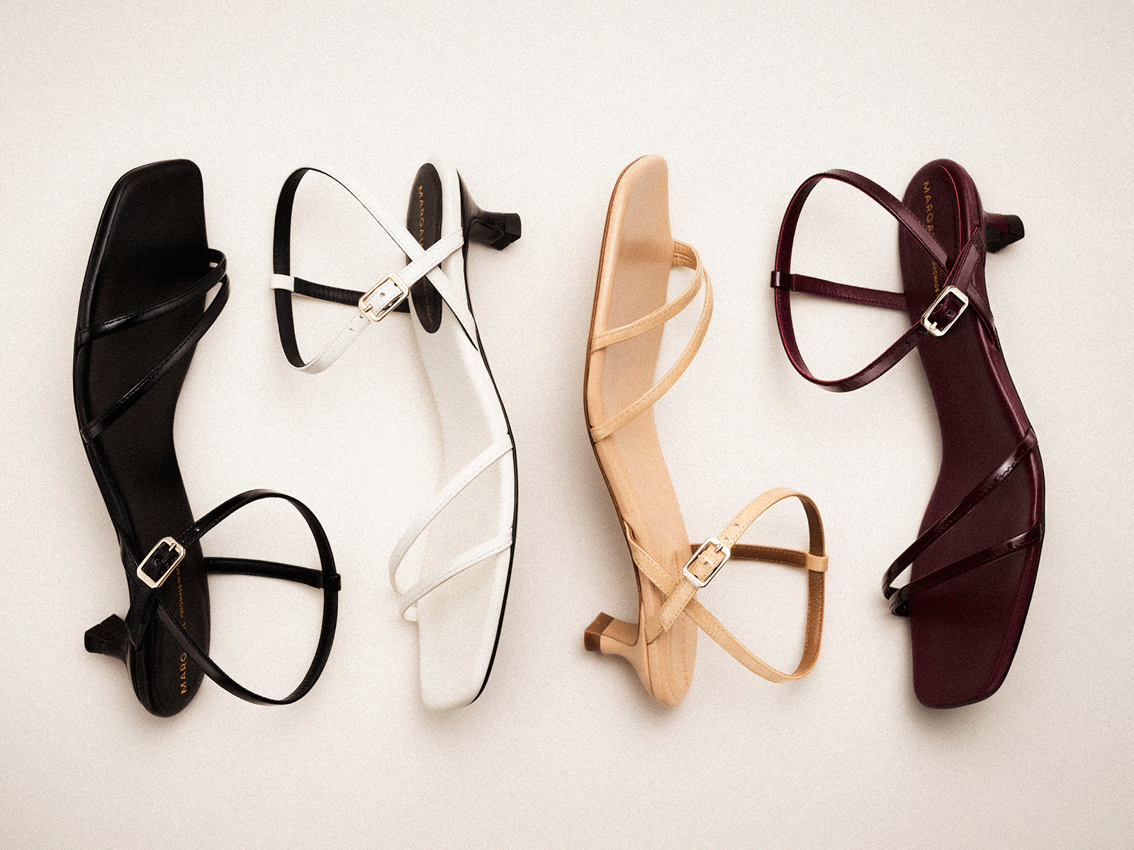Four pairs of strappy heeled sandals in black, white, beige, and burgundy are arranged side by side on a neutral background; each sandal has a low heel and a small buckle.