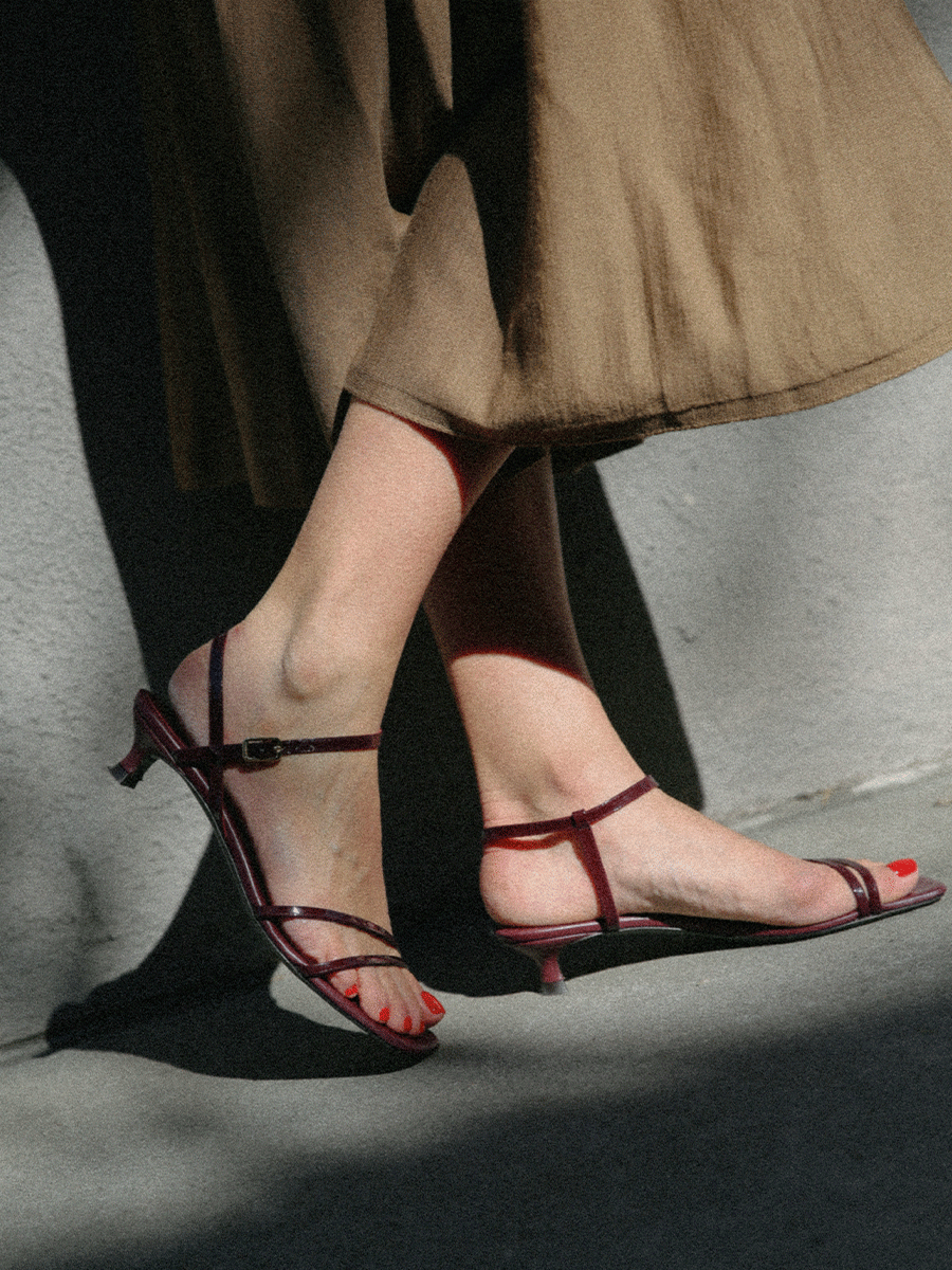 A person wearing a brown skirt and strappy burgundy heeled sandals with red-painted toenails is standing on a sunlit sidewalk. Shadows and light create contrasting patterns on the ground and legs.