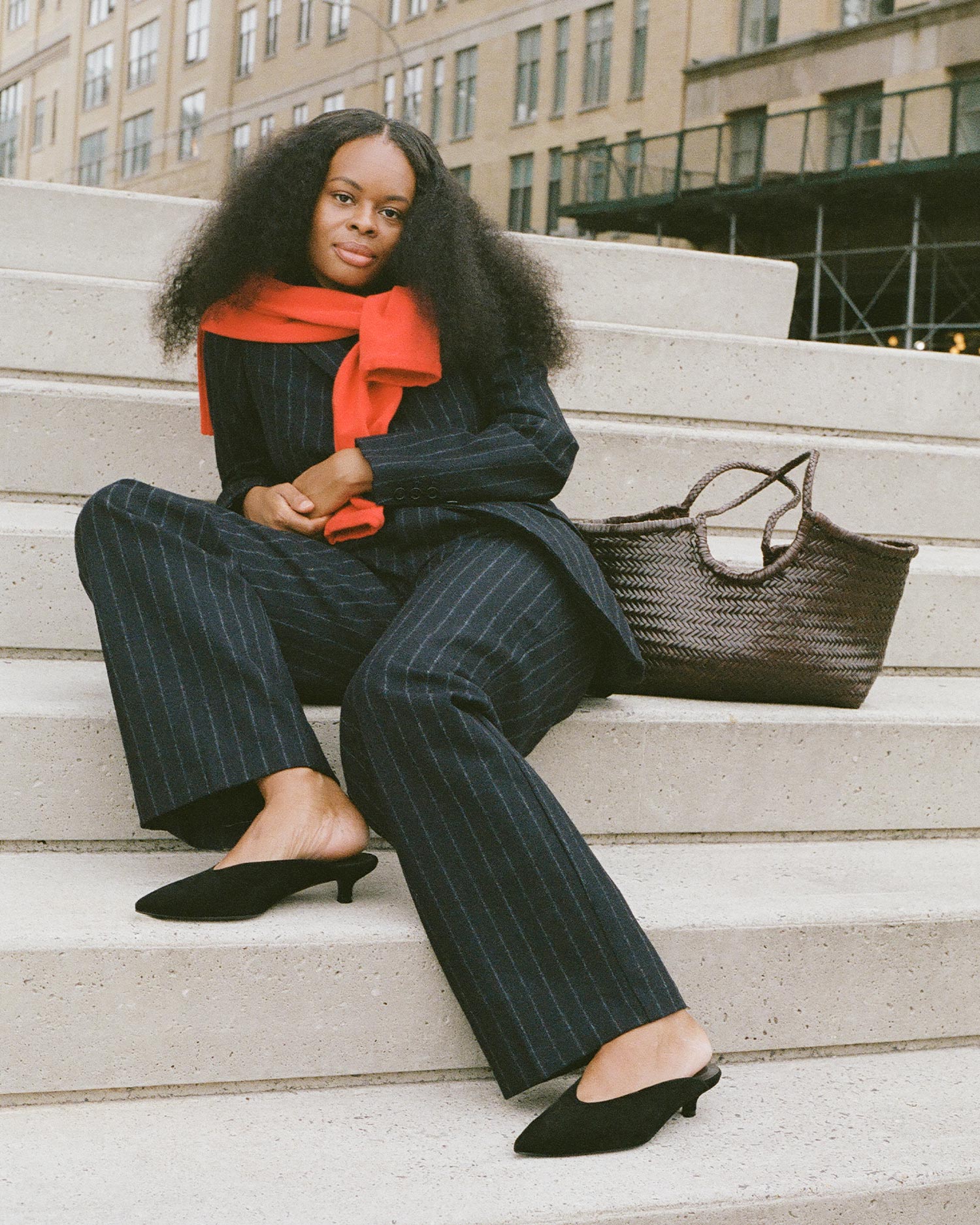 A woman with long, natural hair sits on outdoor steps wearing a dark pinstripe suit, black heels, and a red sweater draped over her shoulders. A large woven handbag rests beside her. Urban buildings appear in the background.