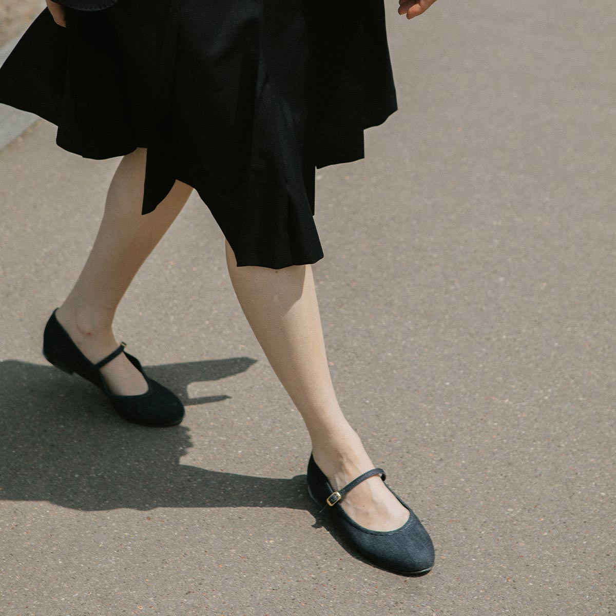 A person exuding elegant femininity walks on a sunlit path, sporting The Demi Jane - Indigo Denim skirt by margaux-development and classic Mary Jane shoes. Only their legs and part of the skirt are visible.