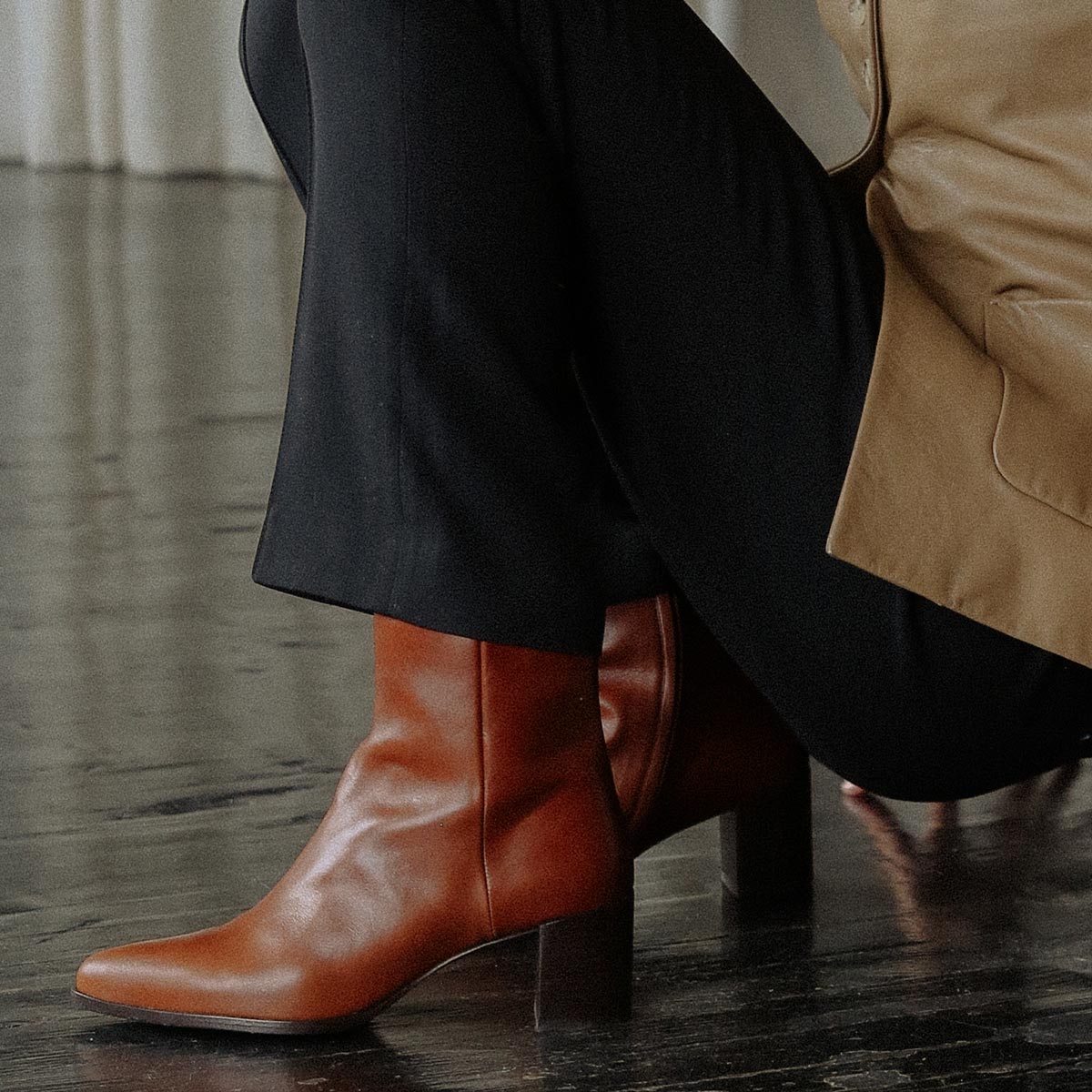 A person in dark pants and a tan ankle-length coat sits on a chair, wearing Margaux’s The Downtown Boot - Saddle Calf; the chic brown leather heeled boots complement the dark wood floor and white curtain backdrop.