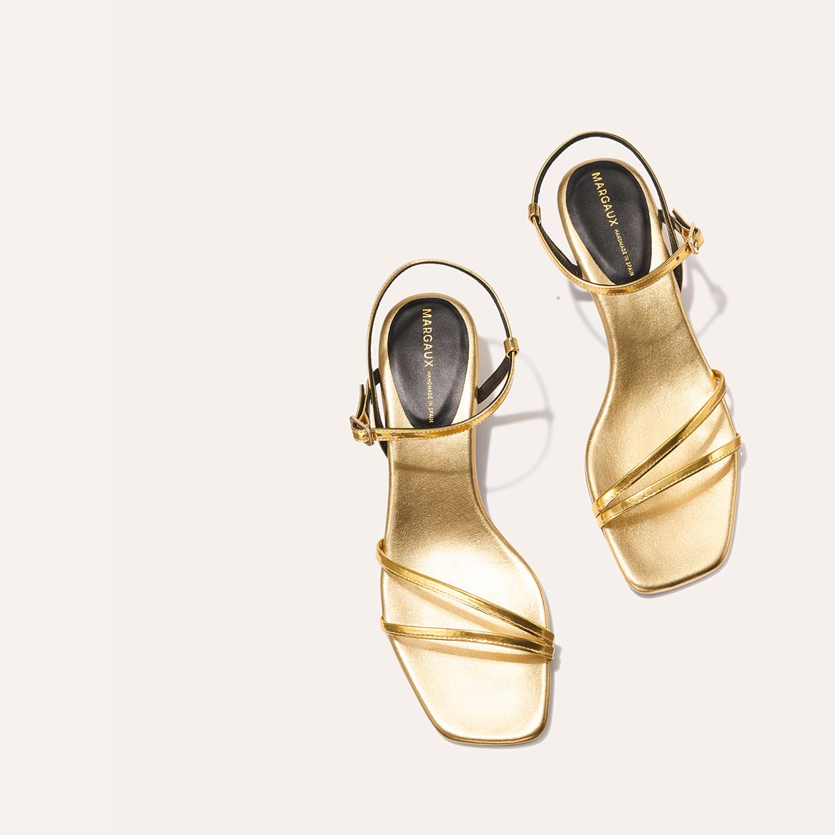 Two gold "The Dylan Sandal - Gold Mirror" high-heeled sandals by margaux-development, featuring square toes, thin ankle straps, and black insoles, are placed on a light beige background.
