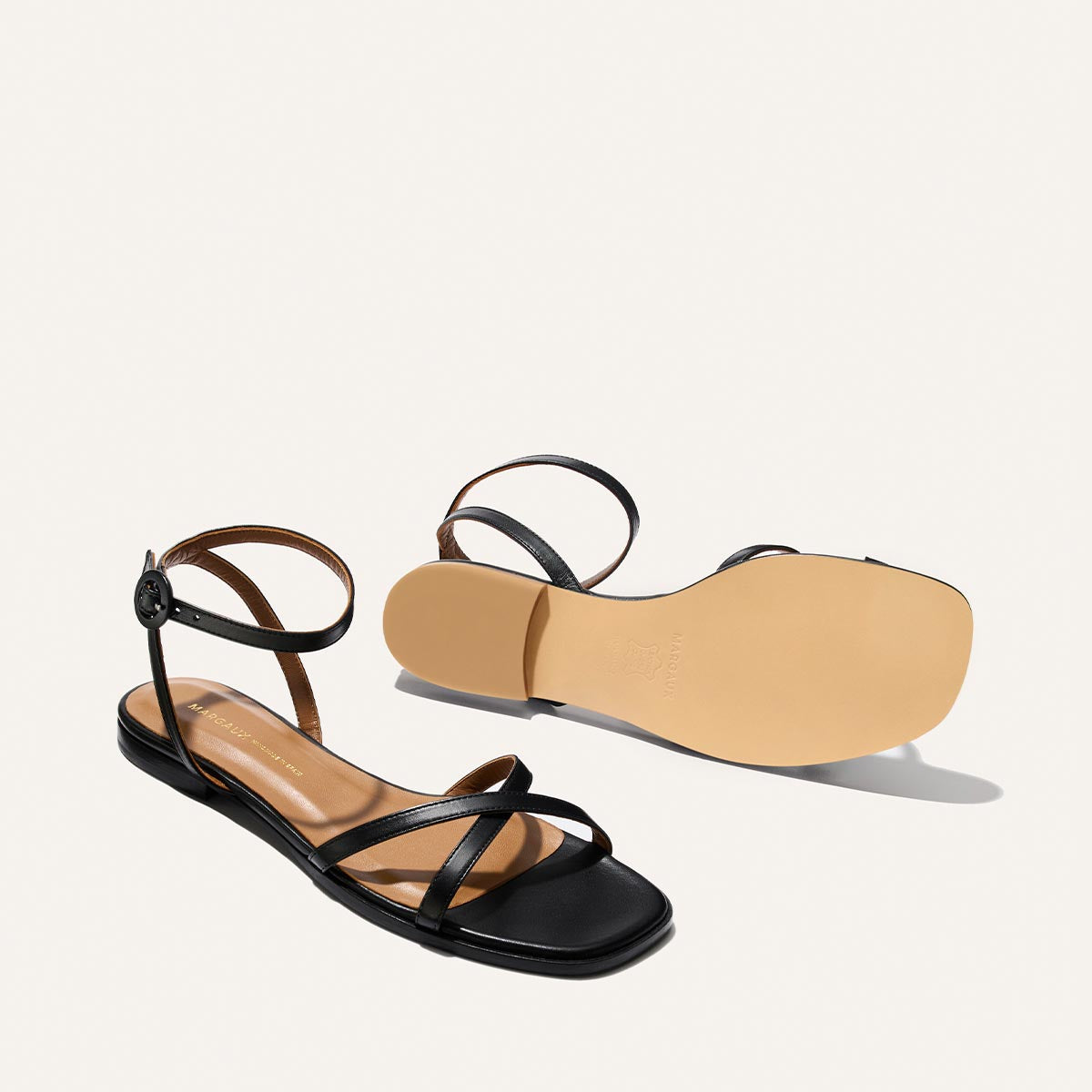 Margaux’s The Flat Sandal – Black Nappa features black strappy flats with ankle straps and buckle closure. One sandal is upright to show the top, the other flipped for the tan sole. Foam-cushioned insoles ensure timeless comfort and style.