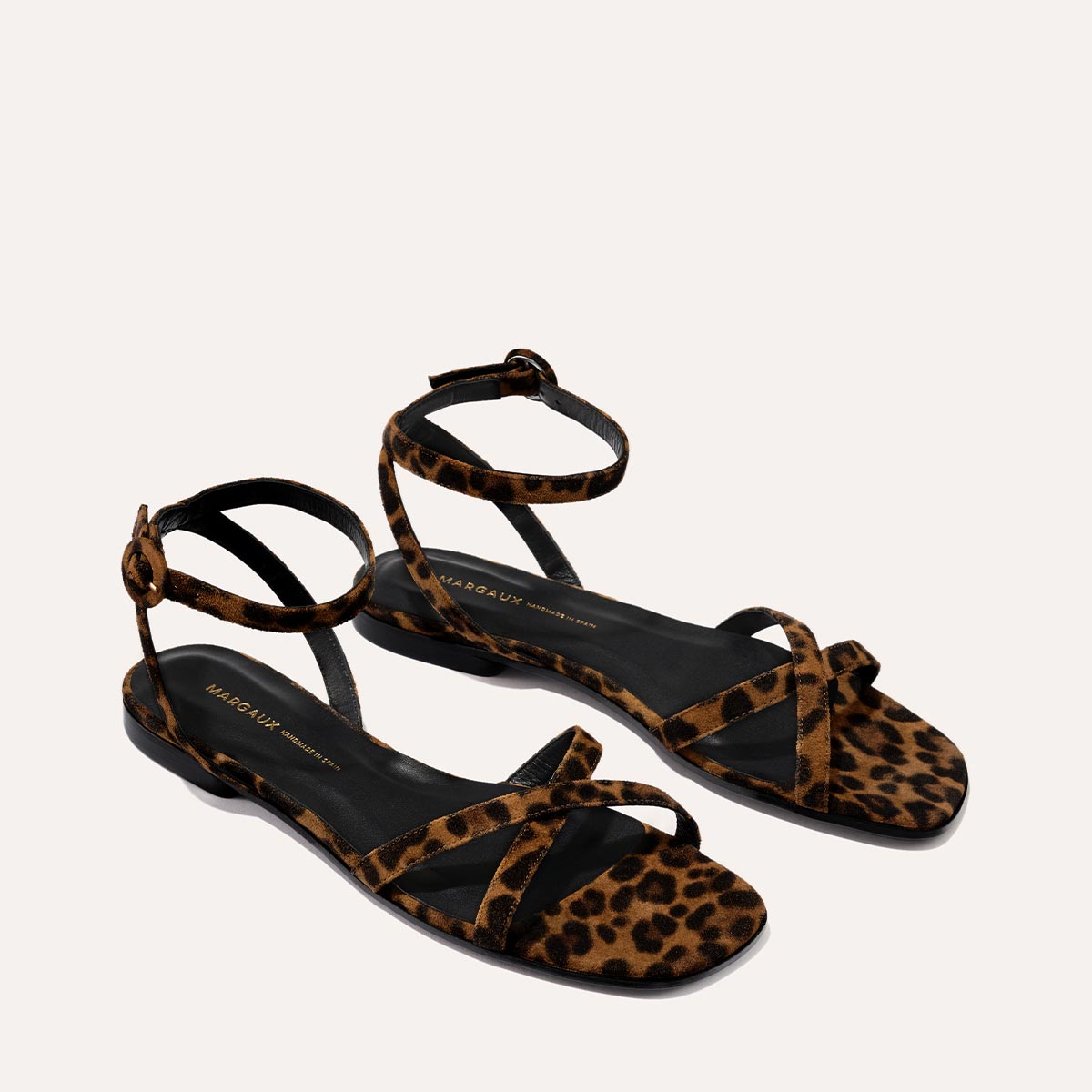The Flat Sandal - Leopard Suede by Margaux: handcrafted womens flat sandals with leopard-print suede straps, ankle buckles, and black soles in a minimalist style, displayed on a light neutral background.