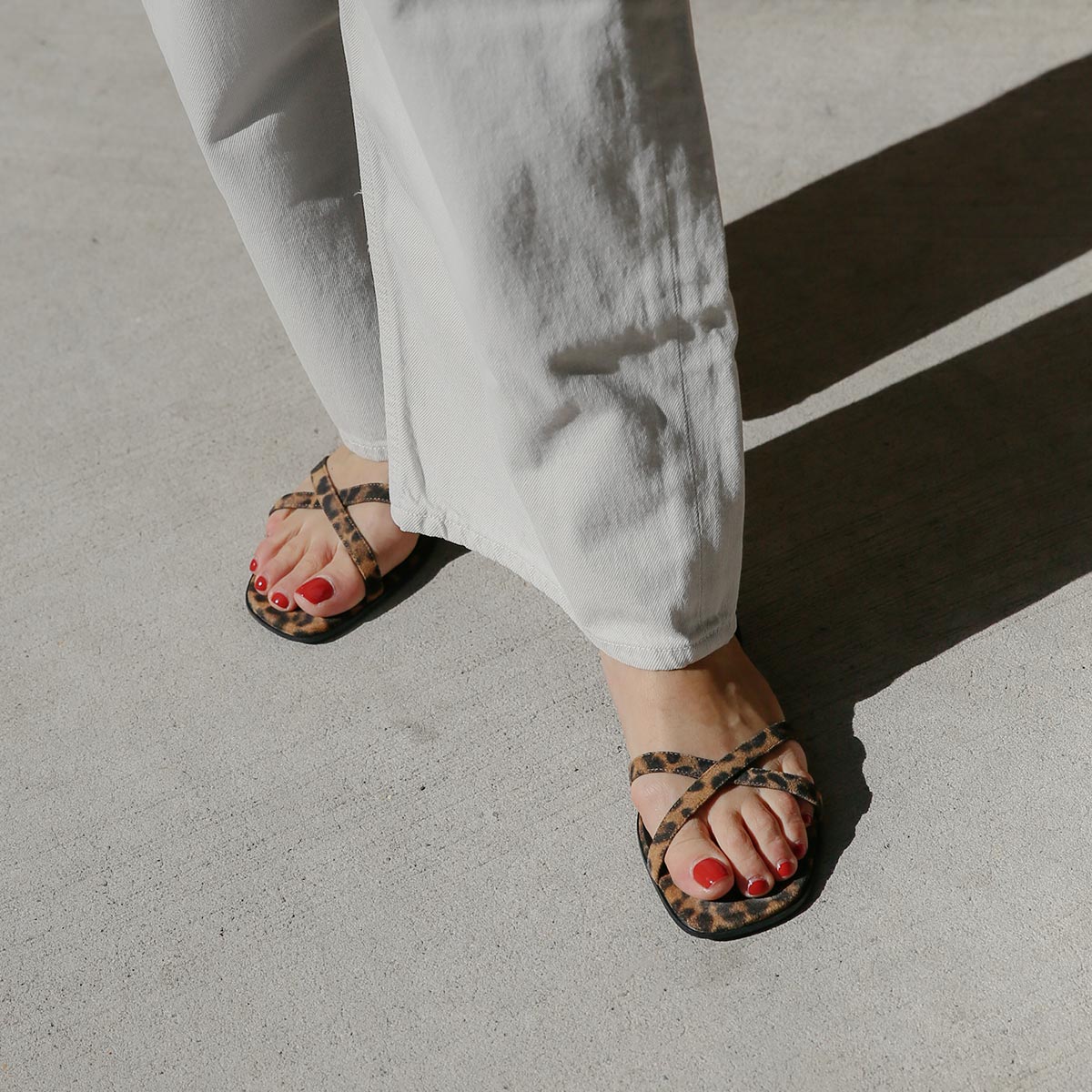 A person wearing white pants and Margauxs The Flat Sandal - Leopard Suede stands on a sunlit concrete surface, showing red-painted toenails.
