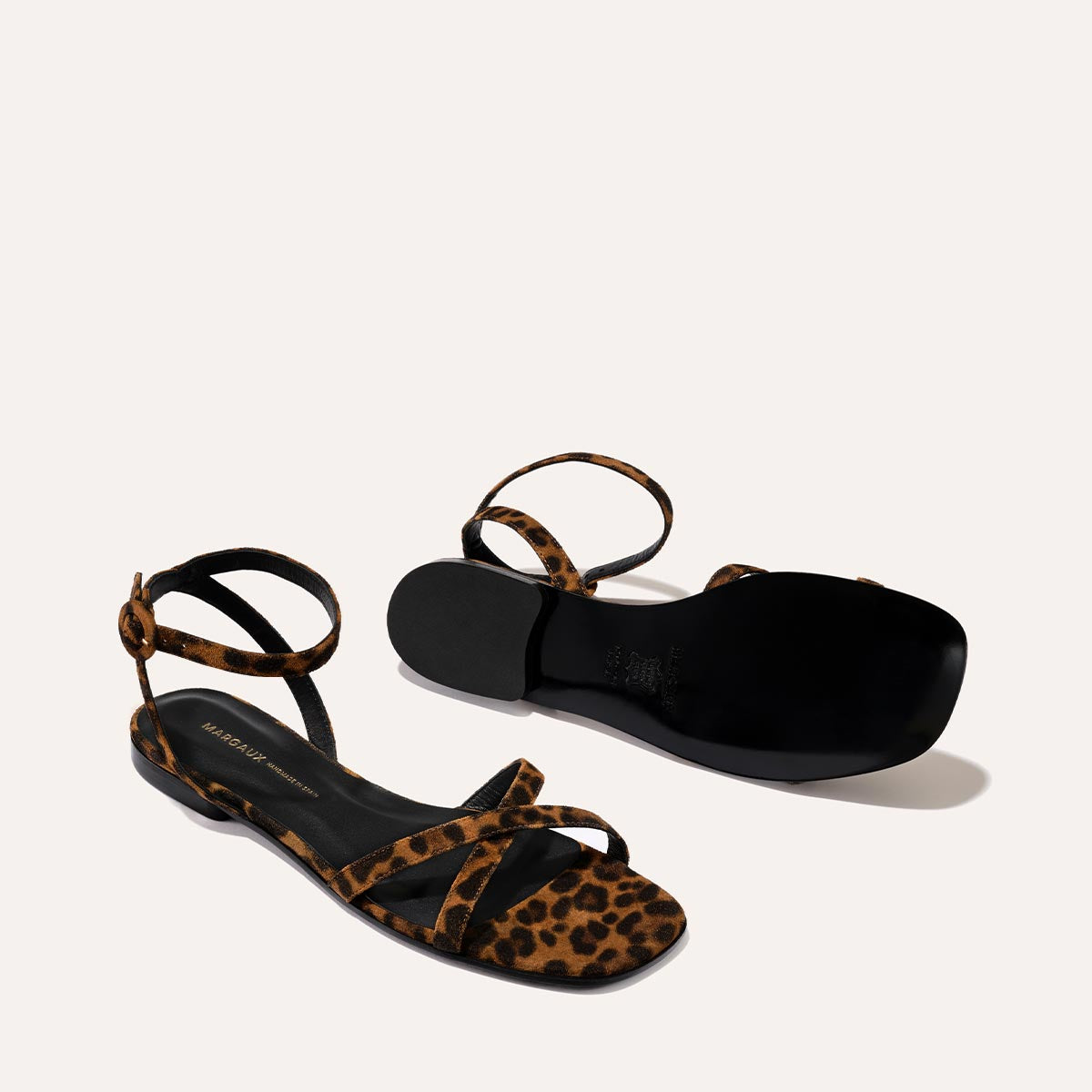 The Flat Sandal - Leopard Suede by Margaux features handcrafted leopard-print suede straps, black insoles, and thin ankle straps. One sandal stands upright while the other is on its side to showcase the black sole.
