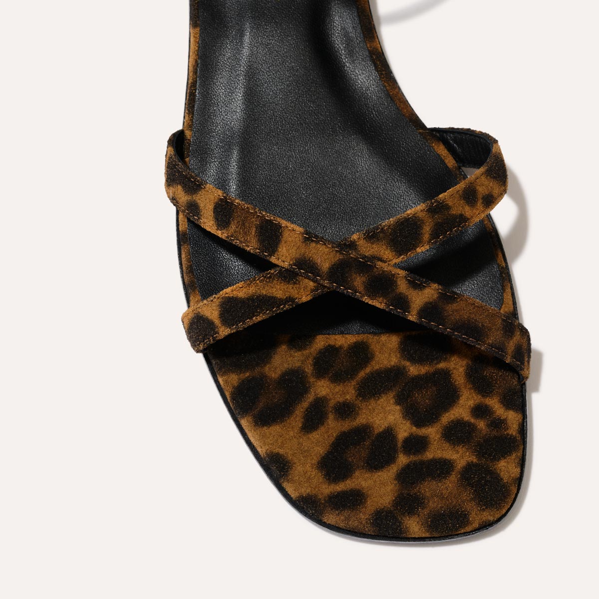 A close-up of Margauxs The Flat Sandal - Leopard Suede, featuring a black sole and crisscross leopard-print suede straps.