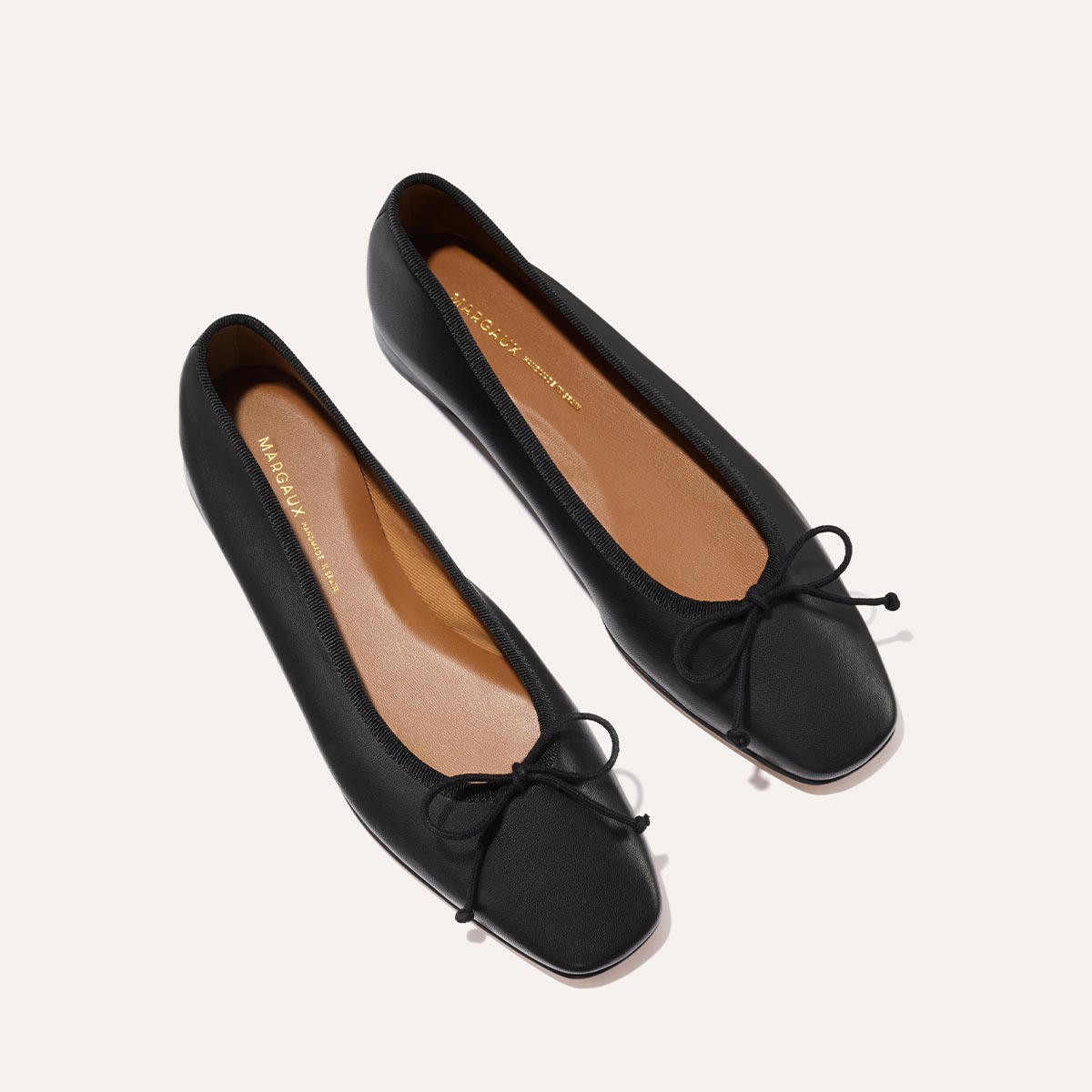 The Fonteyn - Black Nappa by Margaux: handcrafted black ballet flats featuring square toes, petite black bows, tan insoles, and a comfort-first fit, showcased against a light backdrop.