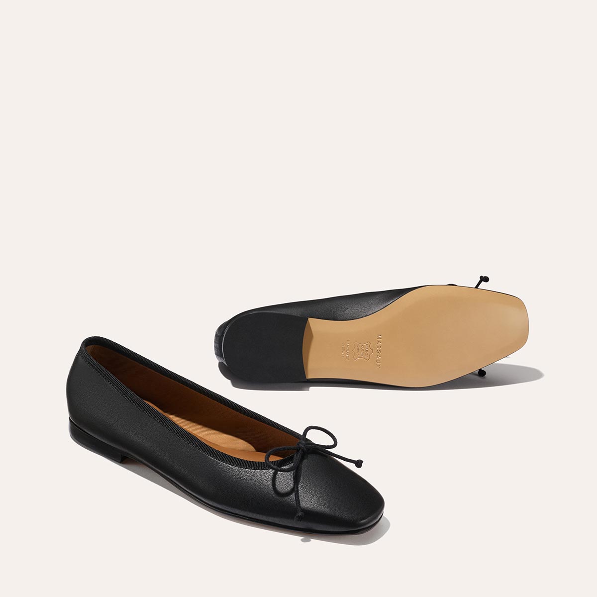 Margaux’s The Fonteyn - Black Nappa: two black leather ballet flats with front bows. One stands upright to show a rounded toe; the other is upside down revealing its tan sole. Both handcrafted shoes rest against a plain light background.