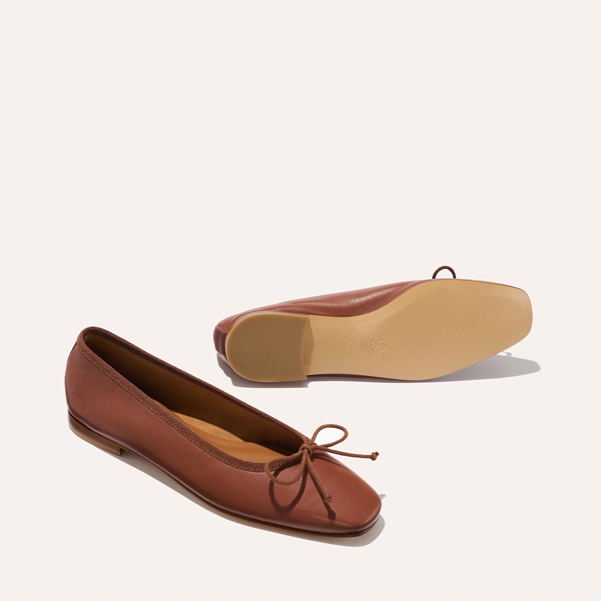 A pair of Margaux’s The Fonteyn ballet flats in Cognac Nappa, featuring a bow on the front; one shoe is upright to show the top and side views, while the other is flipped to reveal the sole, set against a plain light background.