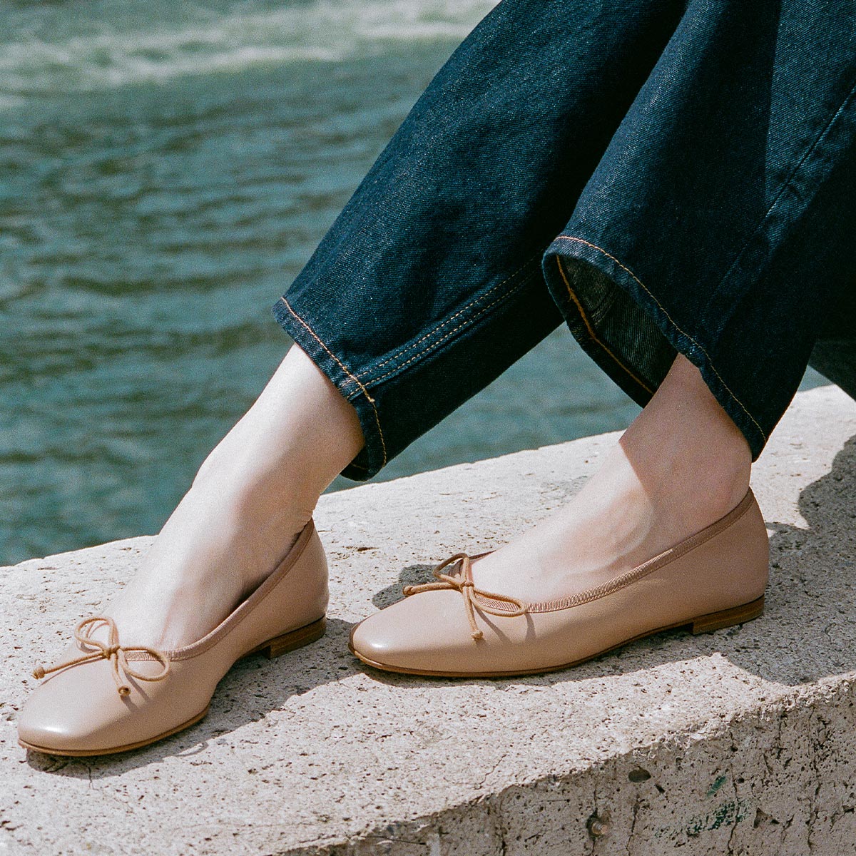 A person in dark blue jeans and Margaux's The Fonteyn ballet flats in Dune Nappa sits on a concrete ledge by the water.