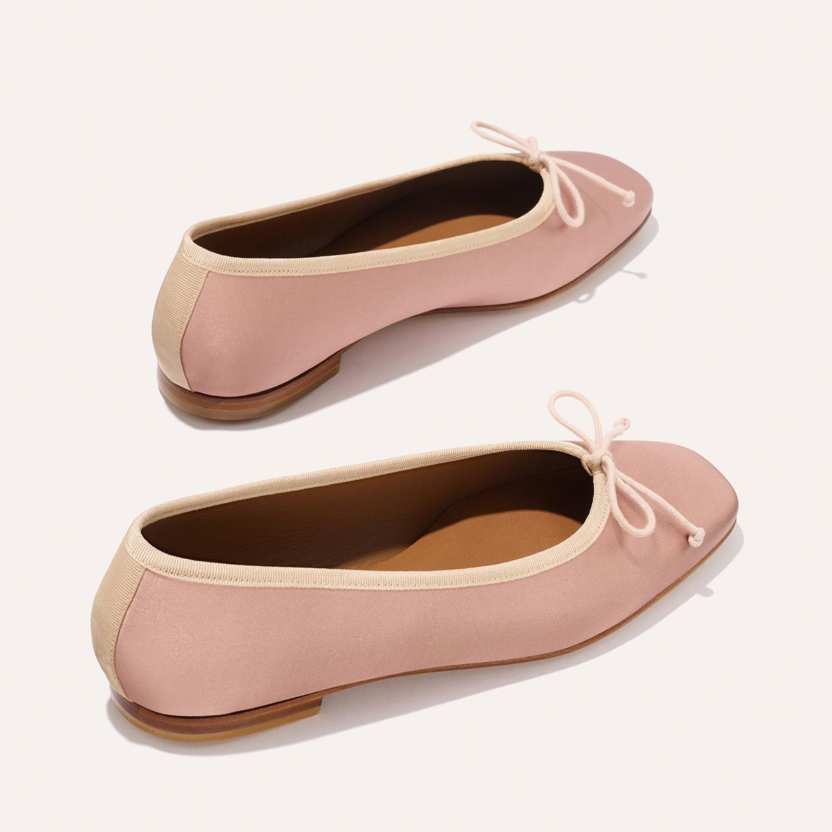 The Fonteyn - Dusty Pink Satin by Margaux: pink ballet flats with tan trim, small front bows, and tan soles, handcrafted for comfort-first fit. Displayed on a light background.