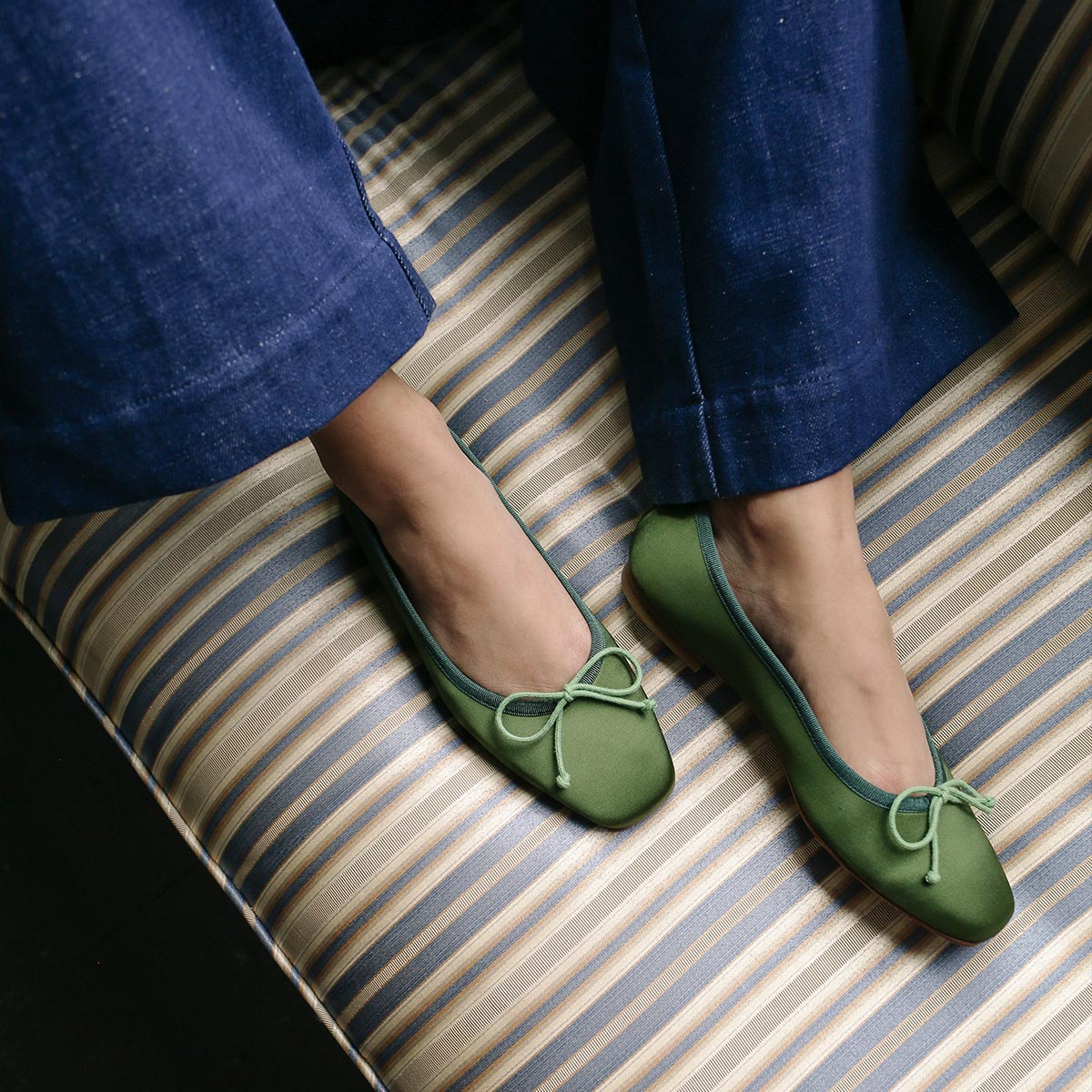 A person in Margaux's The Fonteyn - Forest Satin green ballet flats with bows relaxes on a striped cushioned seat, pairing them stylishly with blue wide-legged pants.