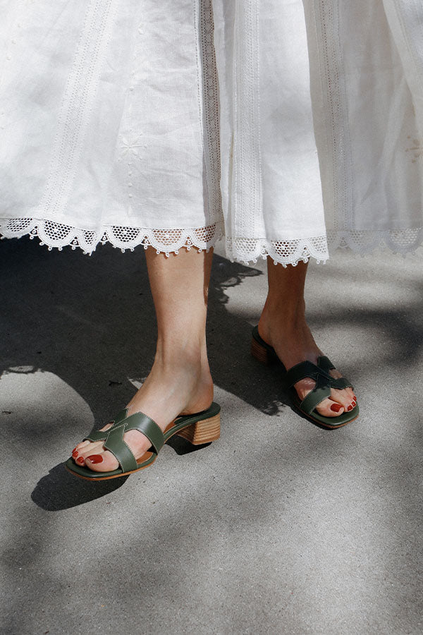 A person wearing a long white skirt with lace trim stands on a sunlit sidewalk, showing green heeled sandals and red painted toenails.