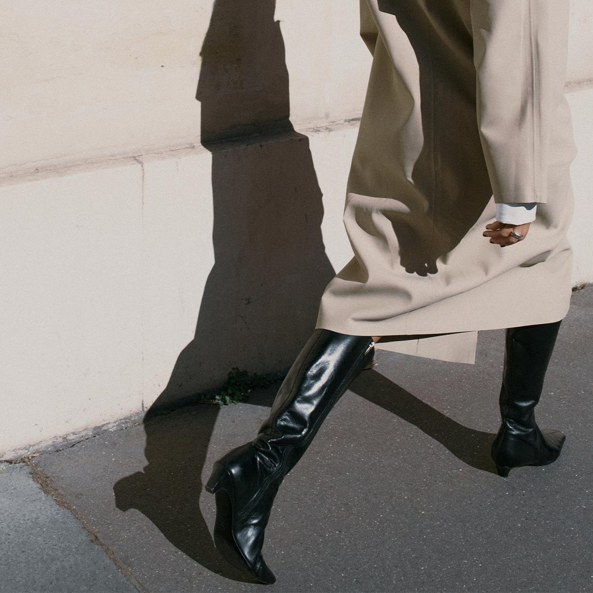 A person walks along a sunlit sidewalk in a long beige coat and Margaux's The Imogen - Black Calf boots, casting a distinct shadow on a pale wall.