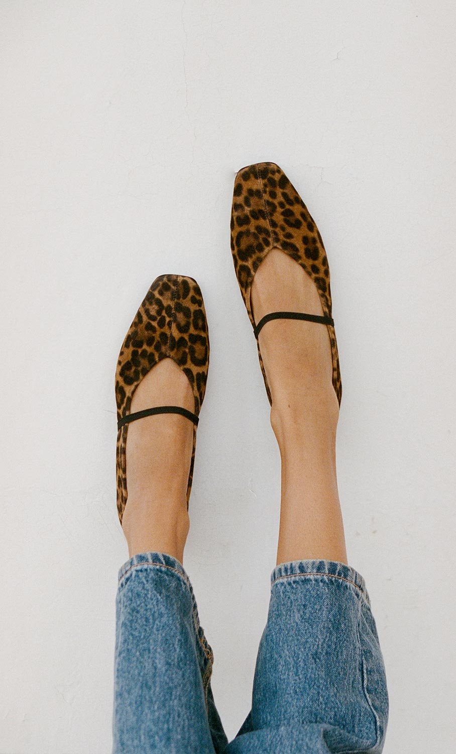 Person wearing blue jeans and leopard print ballet flats with thin black straps, photographed from above against a plain white background.
