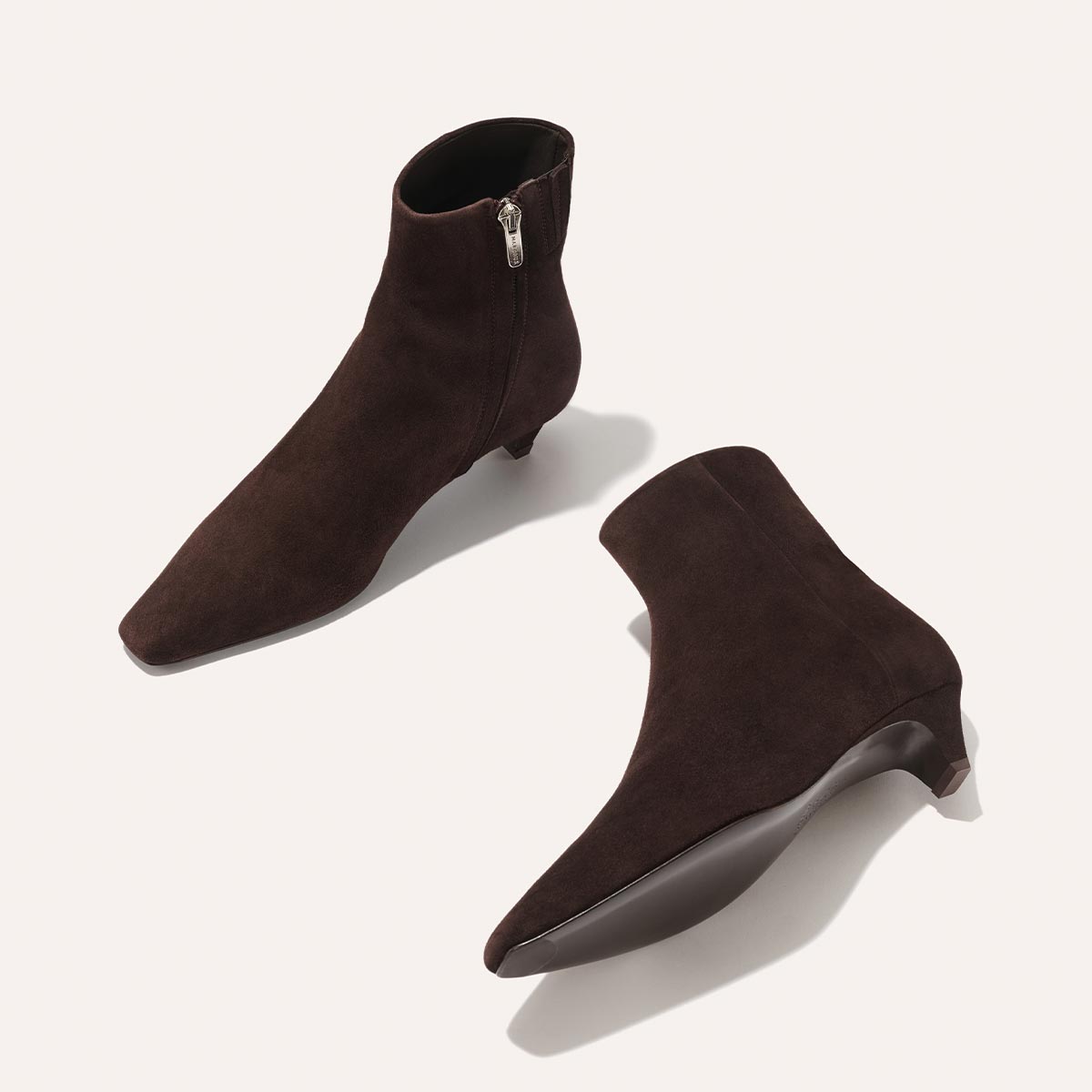 The Alex Mill x Margaux: The Inga - Espresso Suede ankle boots by margaux-development, featuring a low kitten heel, side zipper, and pointed toes, are shown on a light background—one upright and one on its side to reveal the sole.