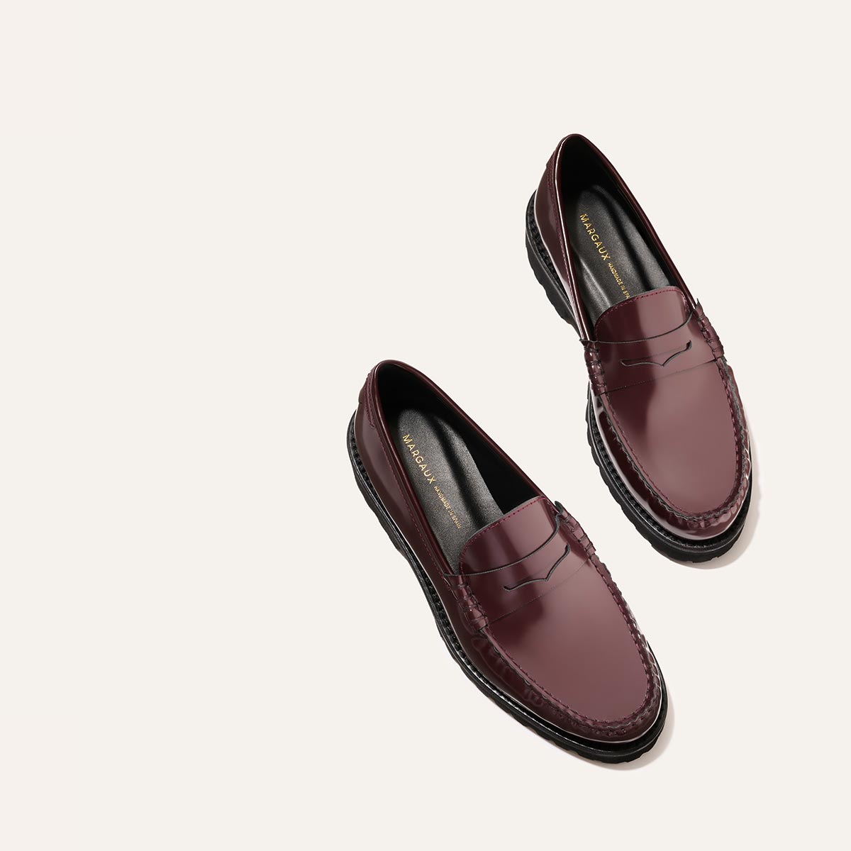 The Louisa Loafer - Burgundy Calf by Margaux, featuring shiny burgundy leather and black lug soles, is displayed diagonally on a light beige background, with the left shoe slightly above the right.