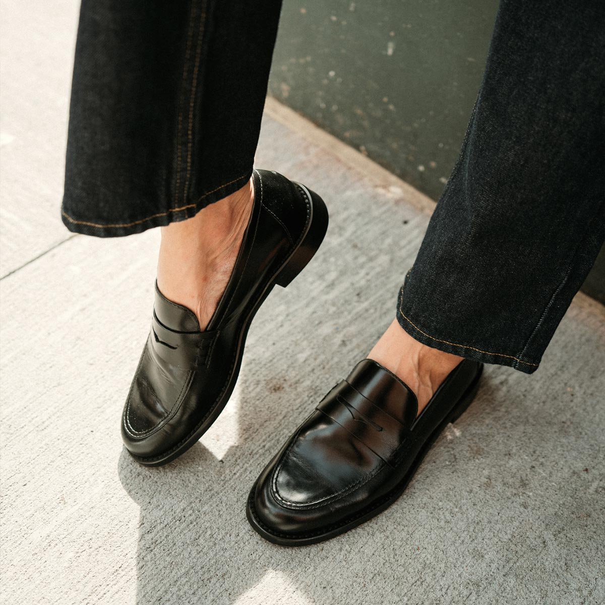 A person wearing margaux-developmentās The Marlowe Loafer in Black Calf and dark jeans stands on a light-colored concrete surface near a green wall.