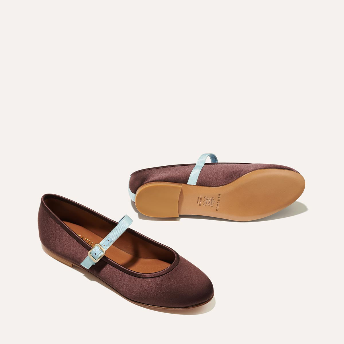 The Demi Jane - Chocolate Satin by Margaux features elegant satin Mary Jane flats with light blue straps and tan soles, shown on a light background—one upright, one on its side—to highlight their refined femininity.