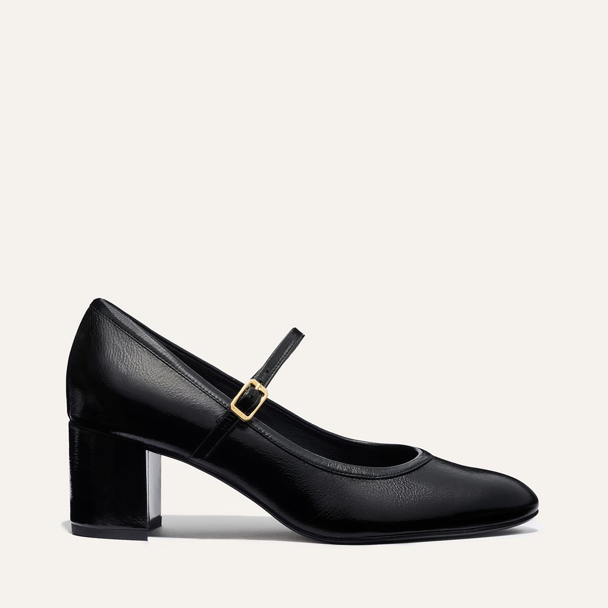The Mira Jane - Black Crinkle Patent by margaux-development is a black crinkle patent leather Mary Jane with a rounded toe, mid-height block heel, and single instep strap secured by a gold buckle.