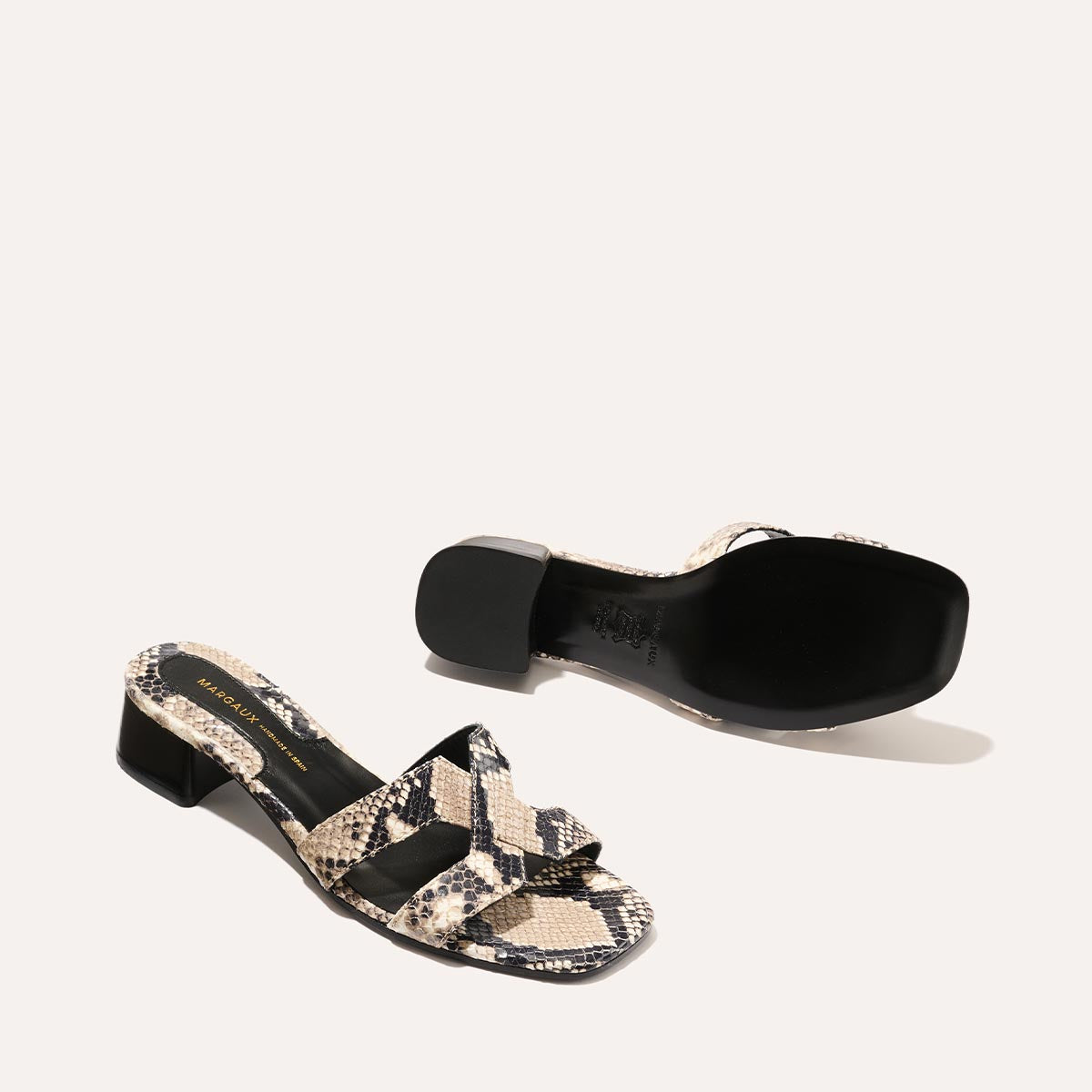 The MX 35 - Natural Python Embossed by margaux-development: two heeled sandals with a black sole and beige, black, and cream faux snakeskin. Both shoes—one upright, one upside down—showcase a block heel on a light background, elevating classic style.