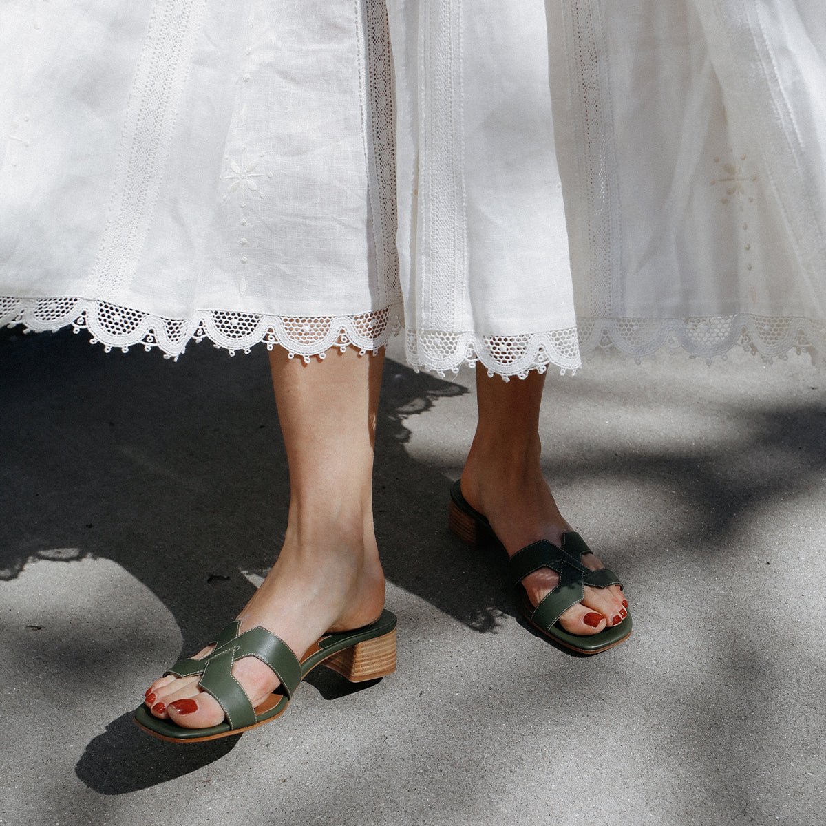 A person wearing a white, lace-trimmed skirt and margaux-developments The MX 35 - Olive Calf block heels stands on a sunlit sidewalk—an elevated take on everyday style.