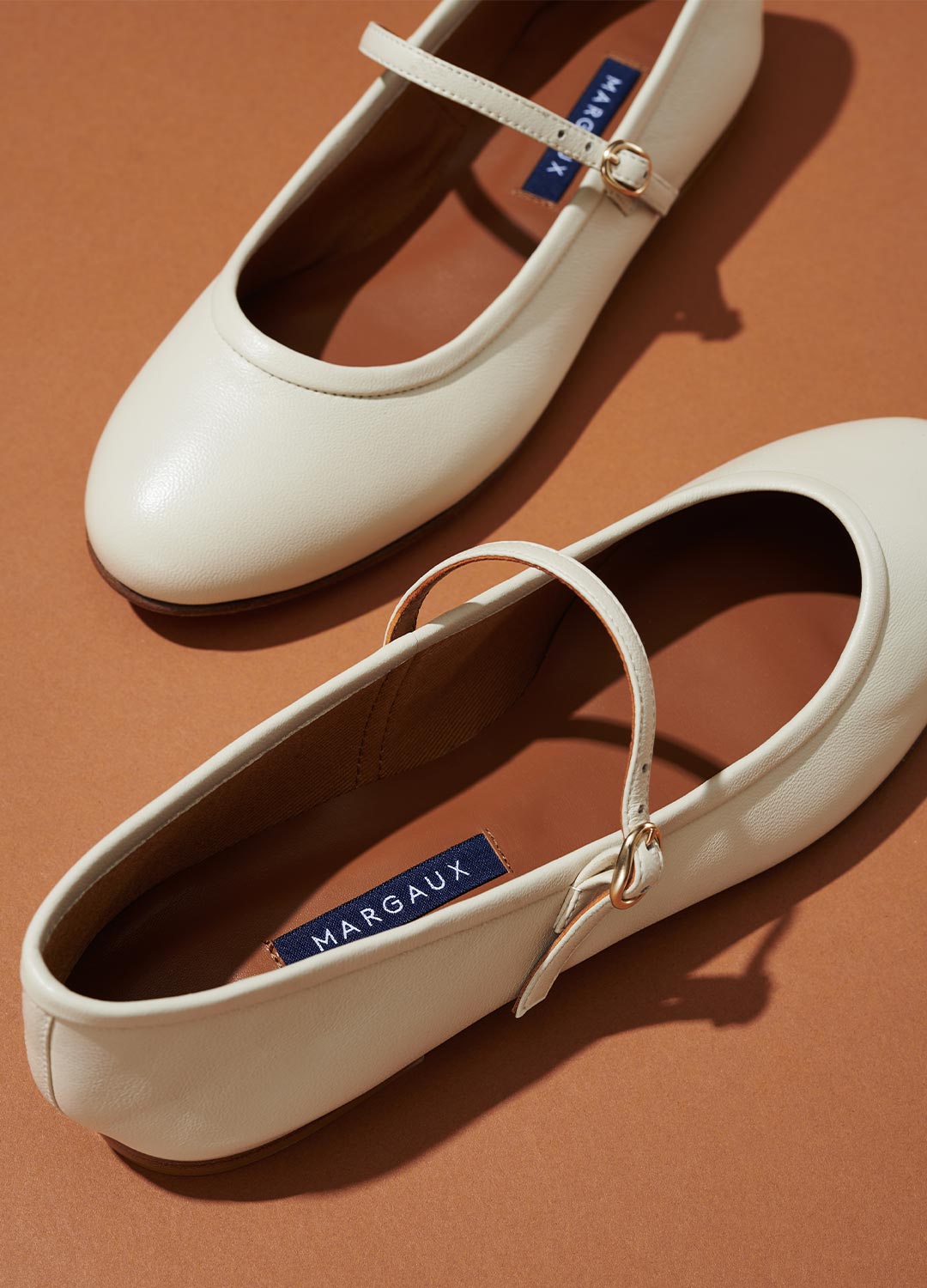 A pair of cream-colored Mary Jane flats with a strap and gold buckle, placed on a brown surface. The label MARGAUX is visible inside the shoes.