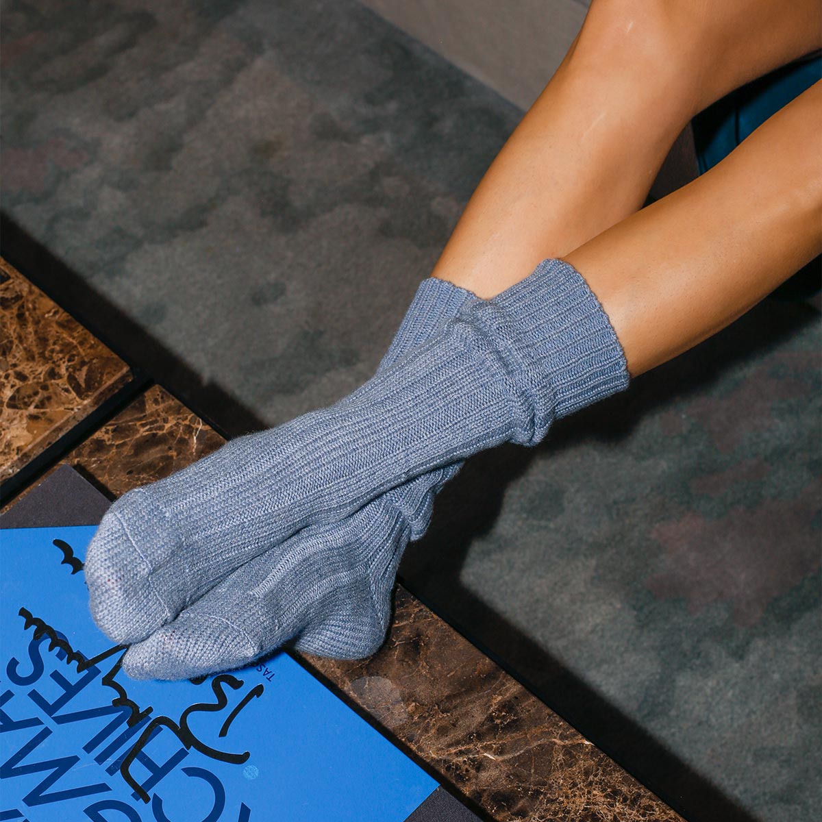 A person with light skin relaxes, feet up on a dark marble table near a blue book, wearing Margaux’s The Cozy Sock in light blue wool/cashmere blend for luxurious comfort.