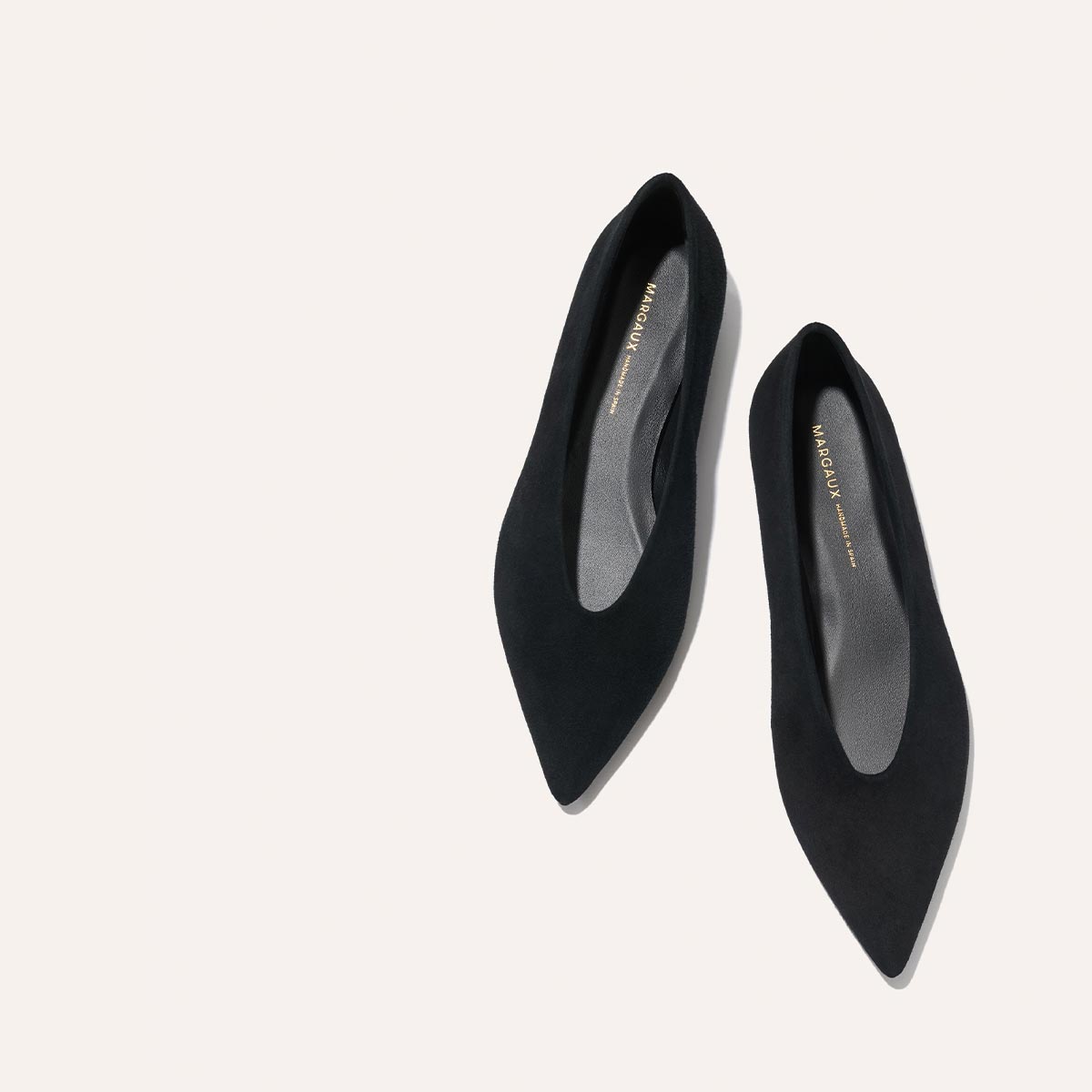 The Parra - Black Suede by margaux-development: black pointed-toe flats with a structured, slightly curved silhouette. Displayed diagonally on a light background, insoles show gold branding.