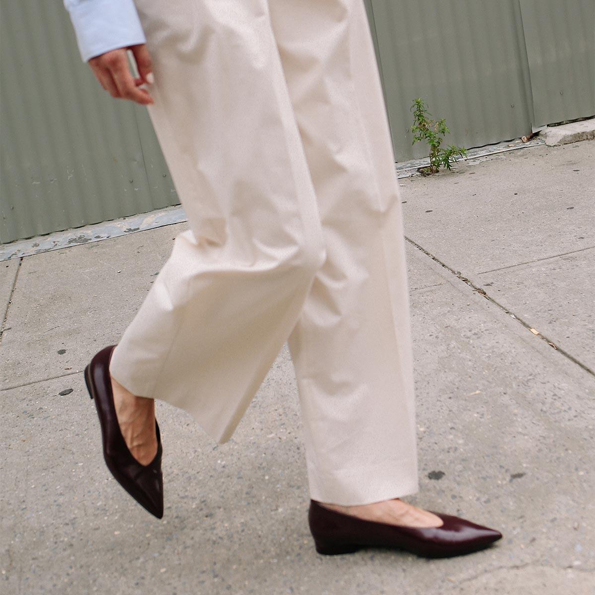 Wearing wide cream pants and The Parra - Burgundy Calf flats by margaux-development, a person walks on a concrete sidewalk with a corrugated metal wall and small weed in the background.