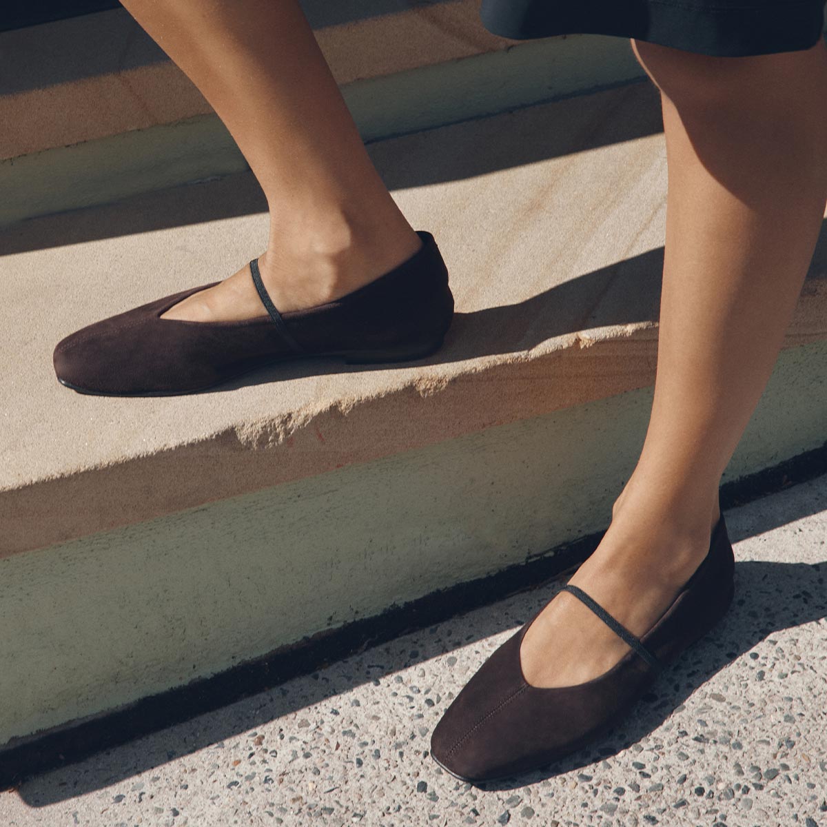 A person wears The Phoebe - Espresso Suede flats by margaux-development, featuring sacchetto construction, as they stand on sunlit outdoor stone steps.