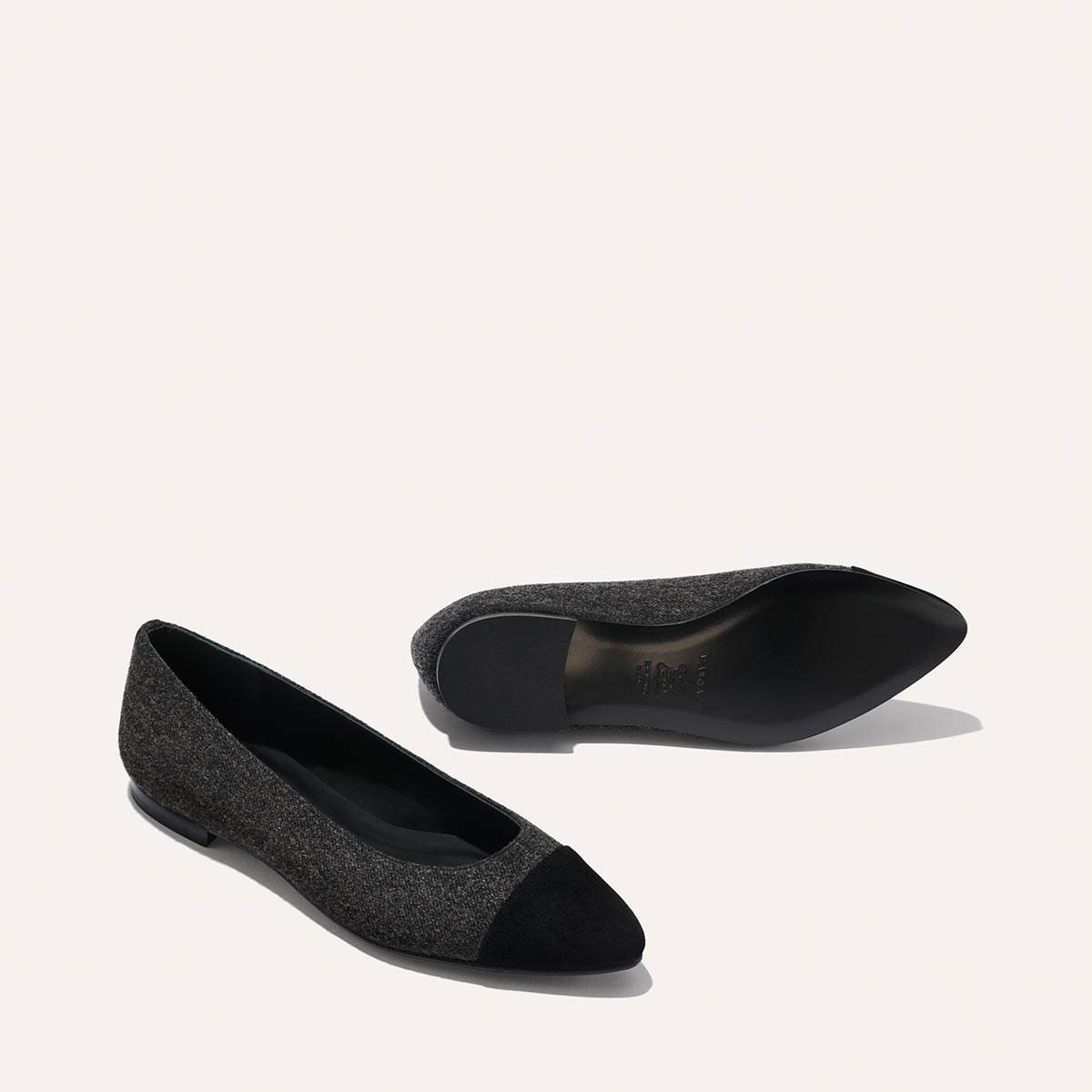Two The Pointe - Grey Flannel flats by margaux-development are displayed on a light background, featuring dark gray uppers with black pointed toe caps. One flat stands upright, while the other lies on its side to reveal the sole.