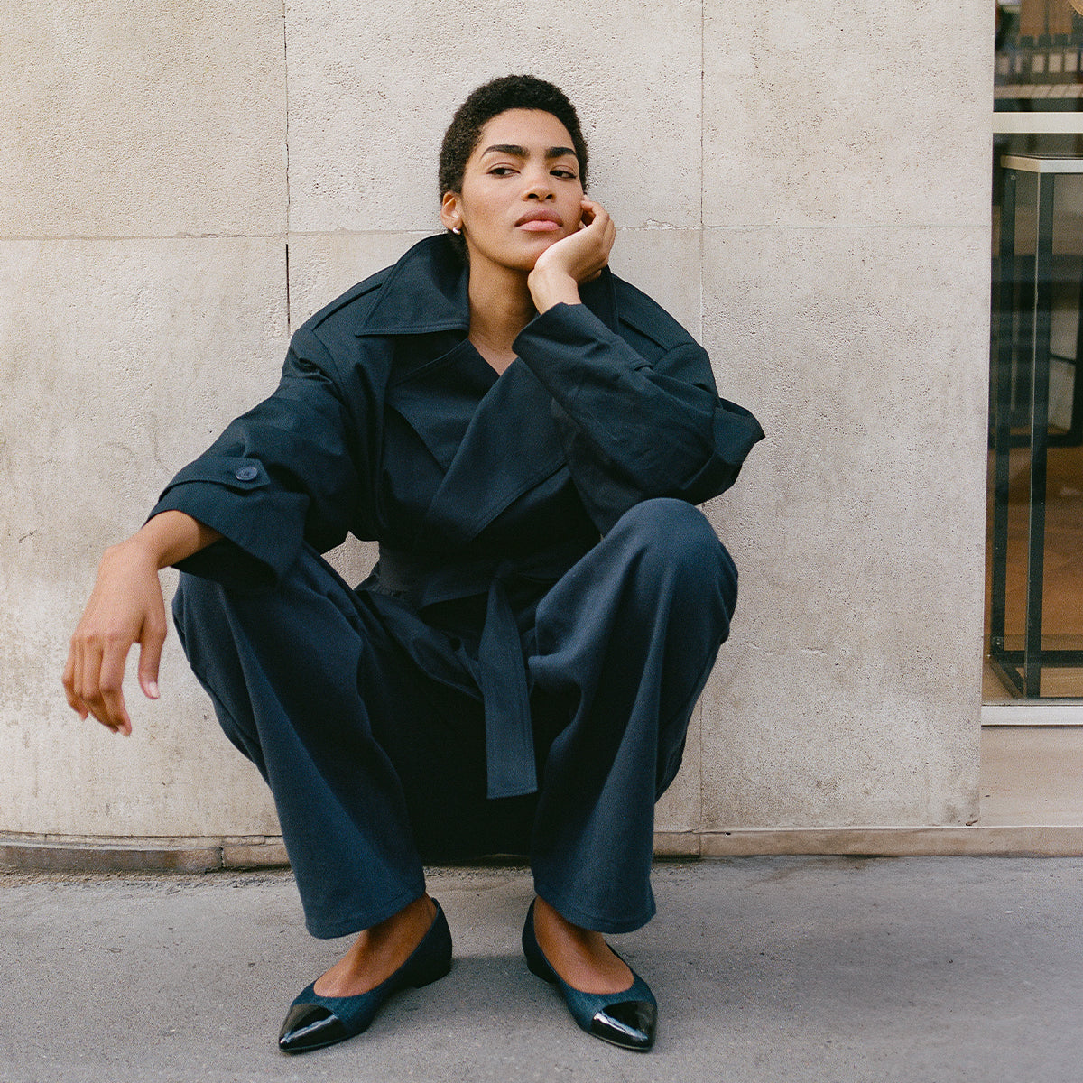 A person wearing The Pointe - Indigo Denim and Patent by margaux-development sits on the sidewalk against a light stone wall, resting their chin on one hand and gazing thoughtfully at the camera.