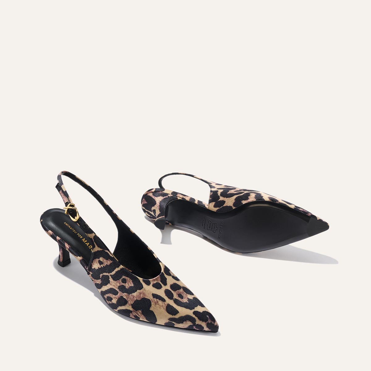 Two The Prado Sling - Leopard Satin heels from margaux-development, featuring pointed toes and mid-height stiletto heels, are shown on a light background—one upright, the other on its side revealing the sole and buckle strap.