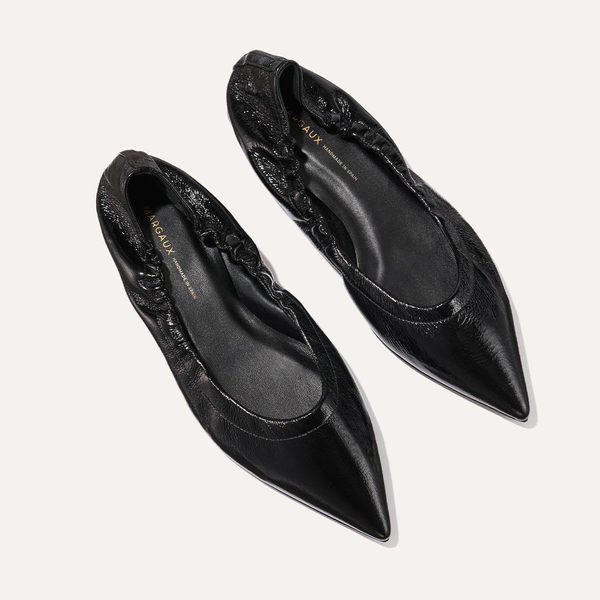 The Margaux The Roma - Black Crinkle Patent features black pointed-toe ballet flats with a gathered, elasticized heel and a smooth patent finish, displayed on a light background.