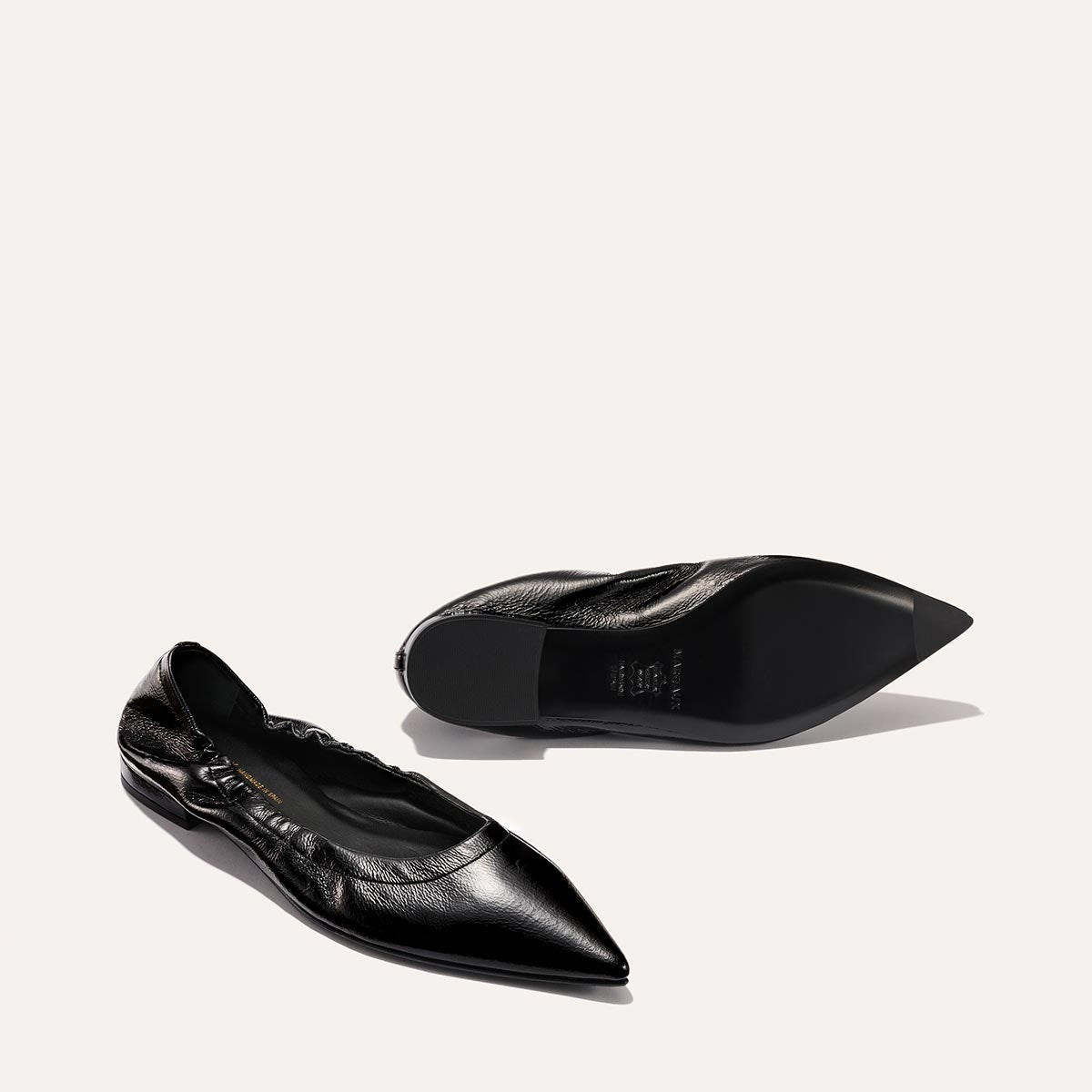 The Roma - Black Crinkle Patent by Margaux: a pair of black pointed-toe ballet flats in textured crinkle patent leather. One stands upright, the other lies on its side to show the sole against a plain, light-colored background.
