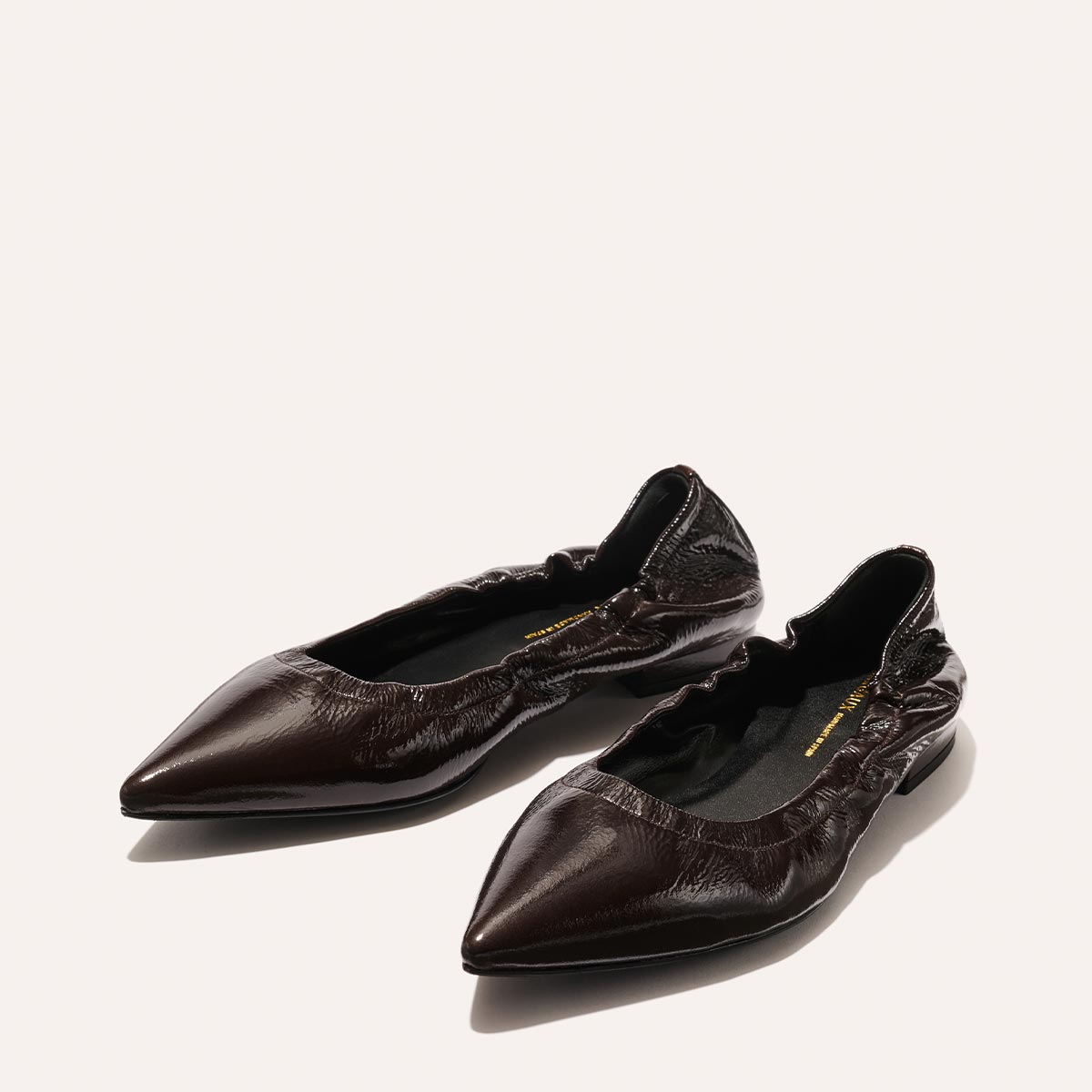 The Roma - Cocoa Crinkle Patent by Margaux are dark brown, pointed-toe ballet flats with a crinkled patent finish and ruched opening, shown on a light background.