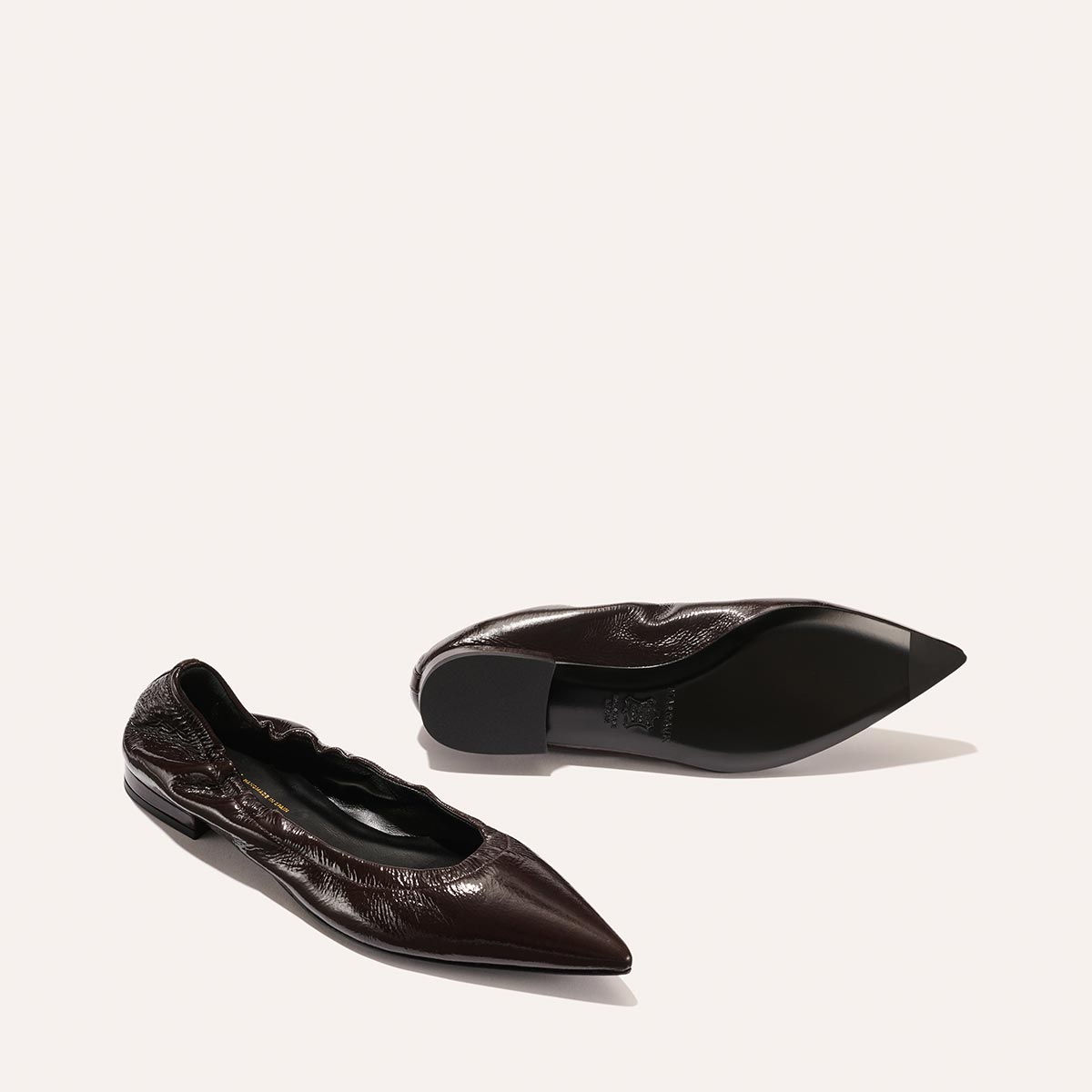 A pair of Margauxs The Roma - Cocoa Crinkle Patent ballet flats, featuring a dark brown, glossy textured finish and pointed toes; one stands upright while the other is upside down to display the sole against a plain background.
