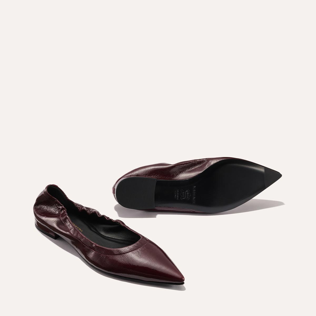 The Roma - Merlot Crinkle Patent ballet flats by Margaux, with pointed toes and burgundy patent leather, are displayed—one upright and one on its side to reveal the sole—against a plain light background, highlighting their sophisticated charm.