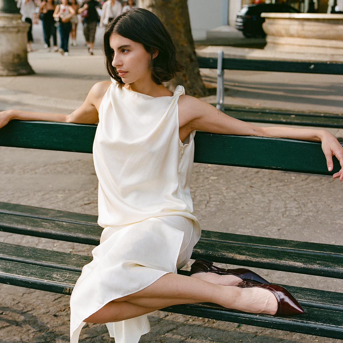 Wearing Margauxs The Roma - Merlot Crinkle Patent flats, a woman in a sleeveless ivory dress sits on a park bench with crossed legs and arms stretched on the backrest, gazing to the side on a sunny day with people and trees in the background.