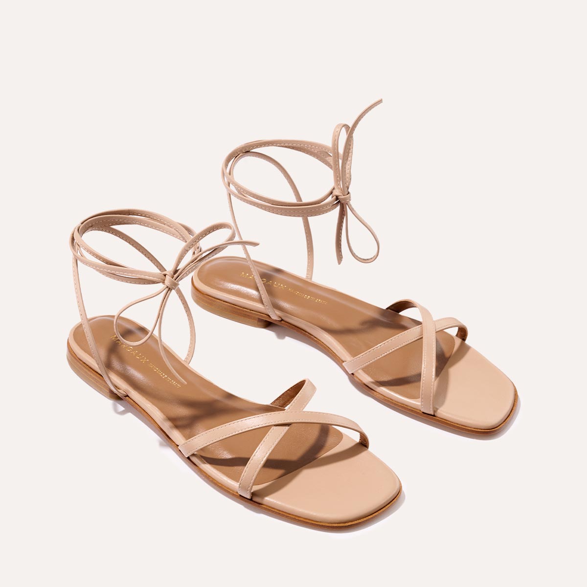 The Wrap Sandal - Rose Nappa by Margaux features a minimalistic design in soft beige with thin ankle ties, offering versatile summertime style. Displayed on a light background.