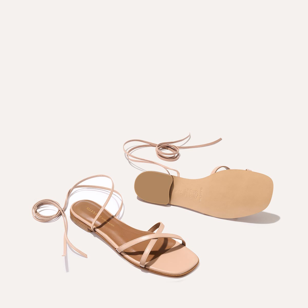 Margauxs The Wrap Sandal in Rose Nappa features nude flat sandals with thin ankle ties—one upright to show the footbed and straps, the other flipped to reveal the sole—capturing their versatile summertime appeal against a pale backdrop.