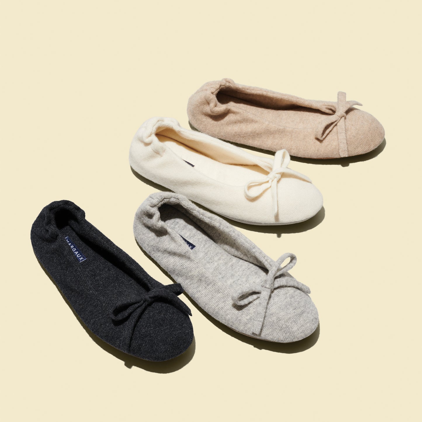 Four pairs of Margaux’s The Ballet Slipper in Oatmeal are arranged diagonally on a light beige background. Each features a cozy wool-cashmere blend and a delicate bow detail, offering the ultimate in stylish comfort.