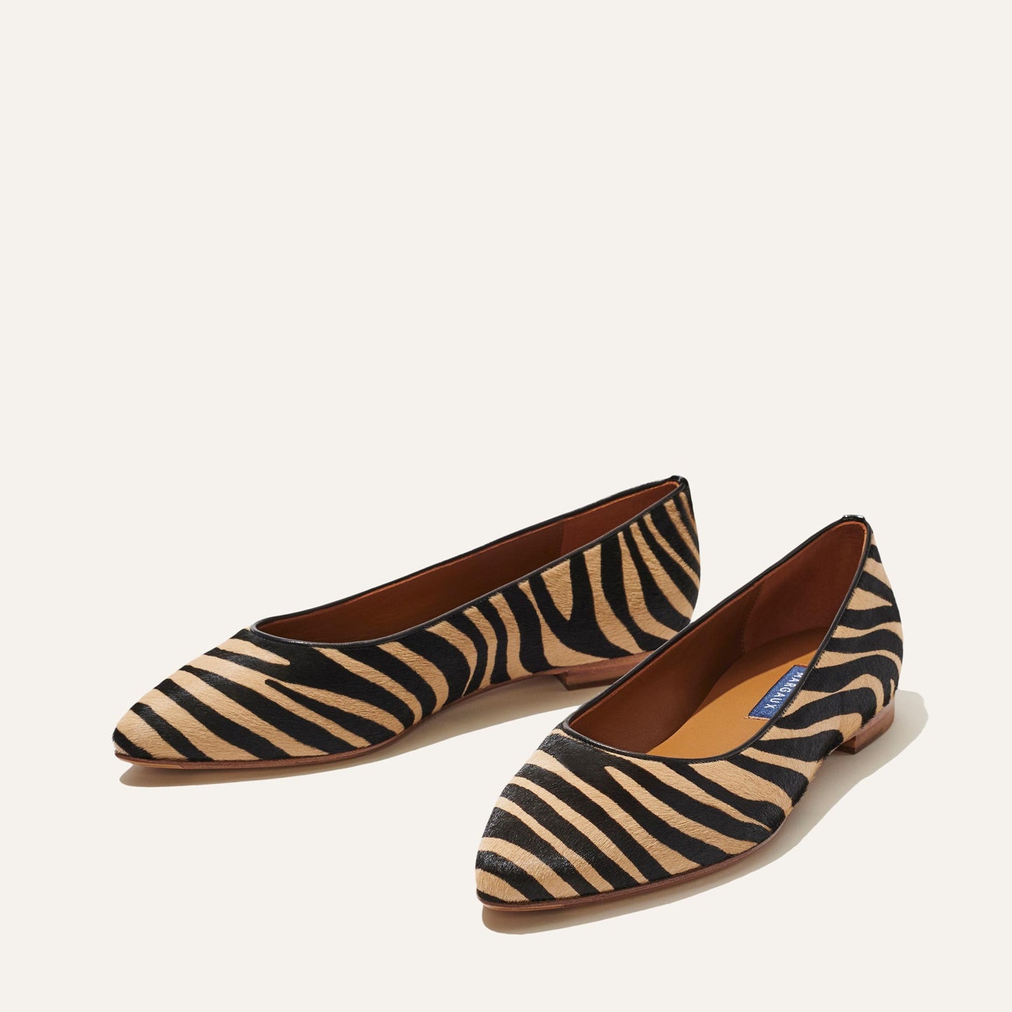 A pair of The Pointe - Zebra Haircalf by Margaux: pointed-toe flats featuring a black and beige zebra stripe, sleek silhouette, low wooden heels, and brown insoles—displayed on an off-white background.