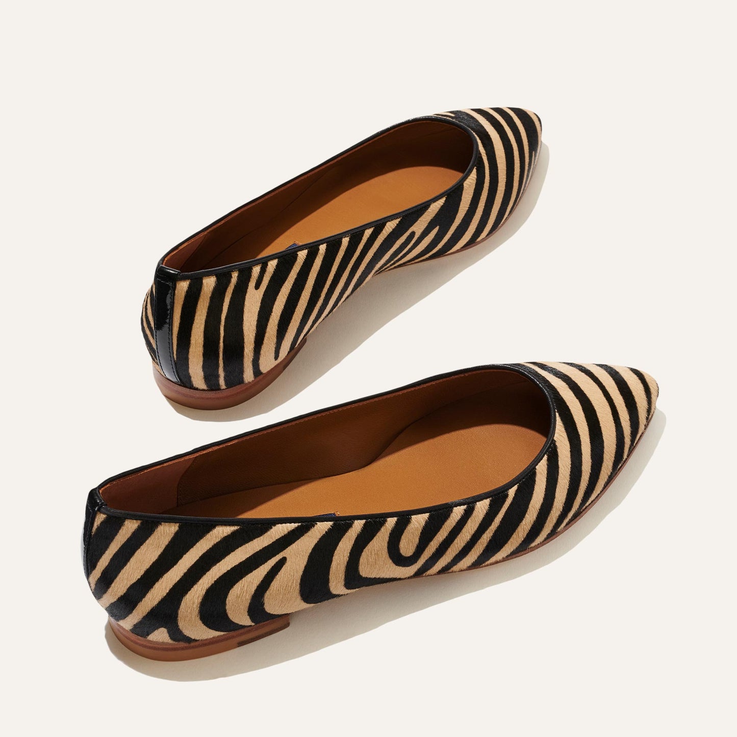 The Margaux The Pointe - Zebra Haircalf flats feature a sleek pointed-toe silhouette with tan and black zebra stripes, brown soles, and tan insoles, displayed on a light background. As seen in Marie Claire.