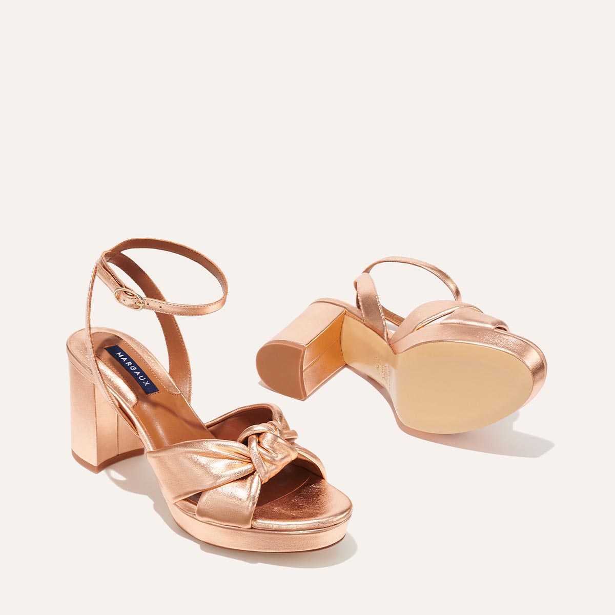 The Carmine Platform - Blush Metallic Nappa by margaux-development features shiny rose gold ankle-strap block heel sandals. One stands upright while the other lies on its side, displaying the sole for a stylish and comfortable look.