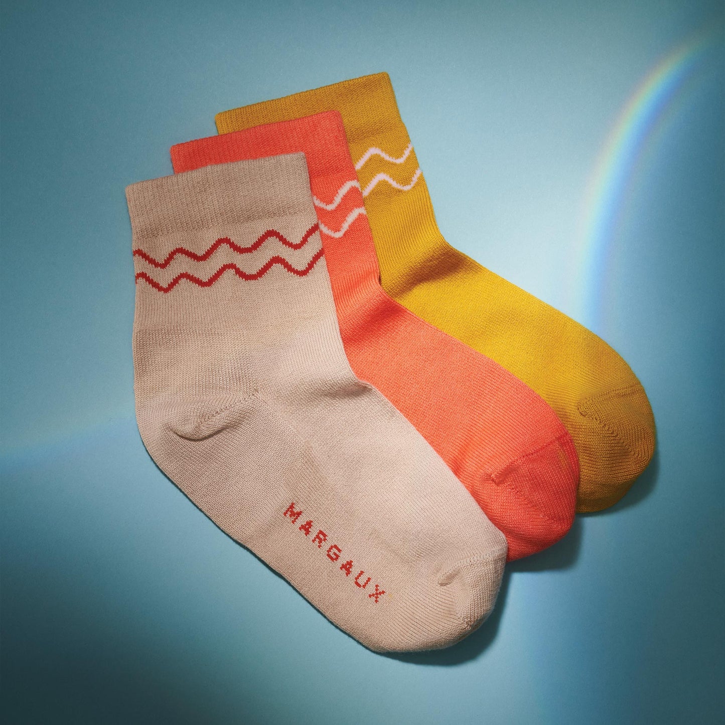 Three pairs of Margaux's The Boot Sock in Camel, orange, and yellow with wavy line patterns near the top. The beige pair features "MARGAUX" on the foot. Socks are overlapped on a blue background.